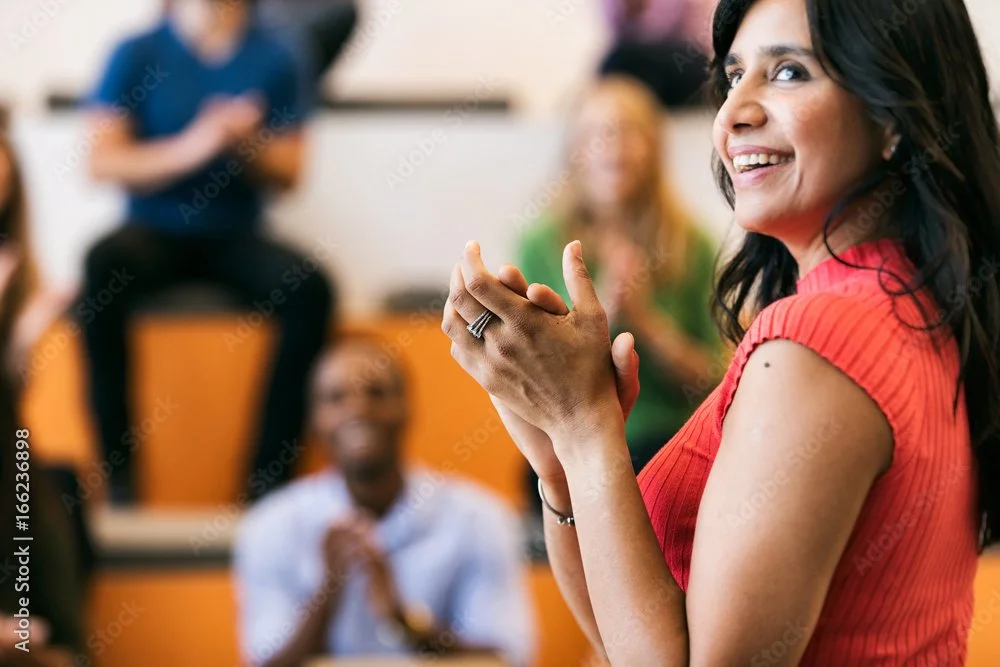 Smiling woman in a red top claps her hands in a group setting with others in the background.