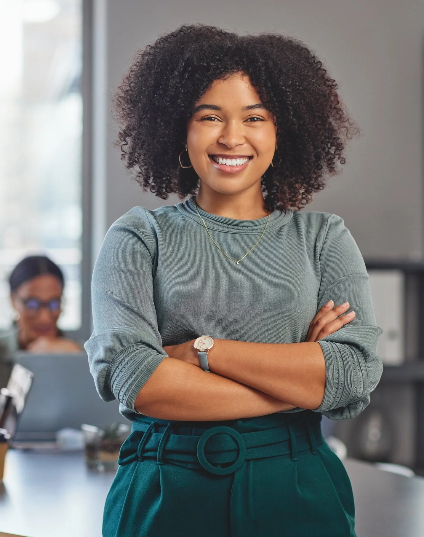 A smiling woman with curly hair crossing her arms in an office setting, wearing a gray long-sleeve top, teal pants with a belt, a silver watch, and a necklace, with colleagues working in the background.