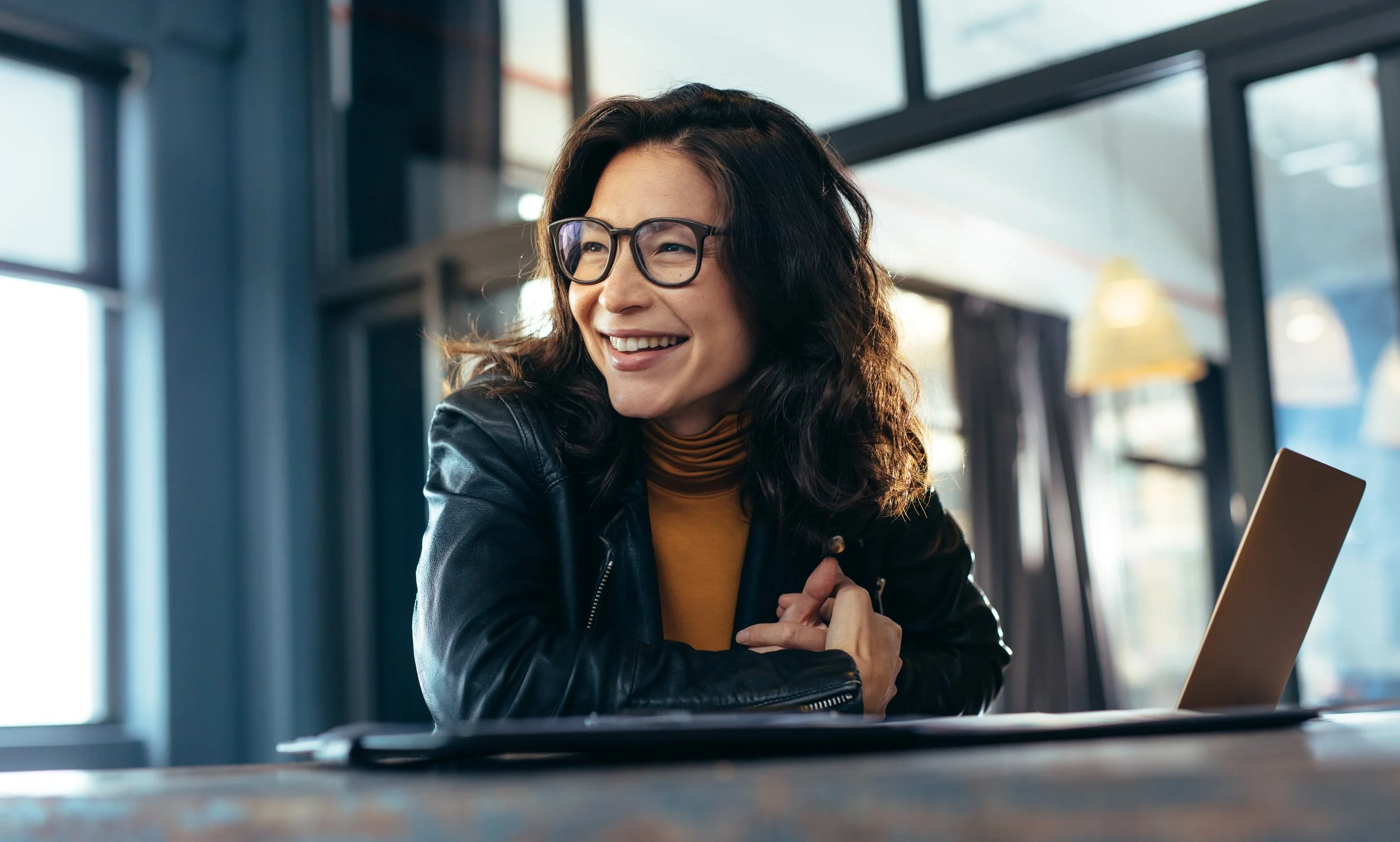 A woman with glasses and curly hair smiling while sitting at a table with a laptop in a modern, well-lit cafe.