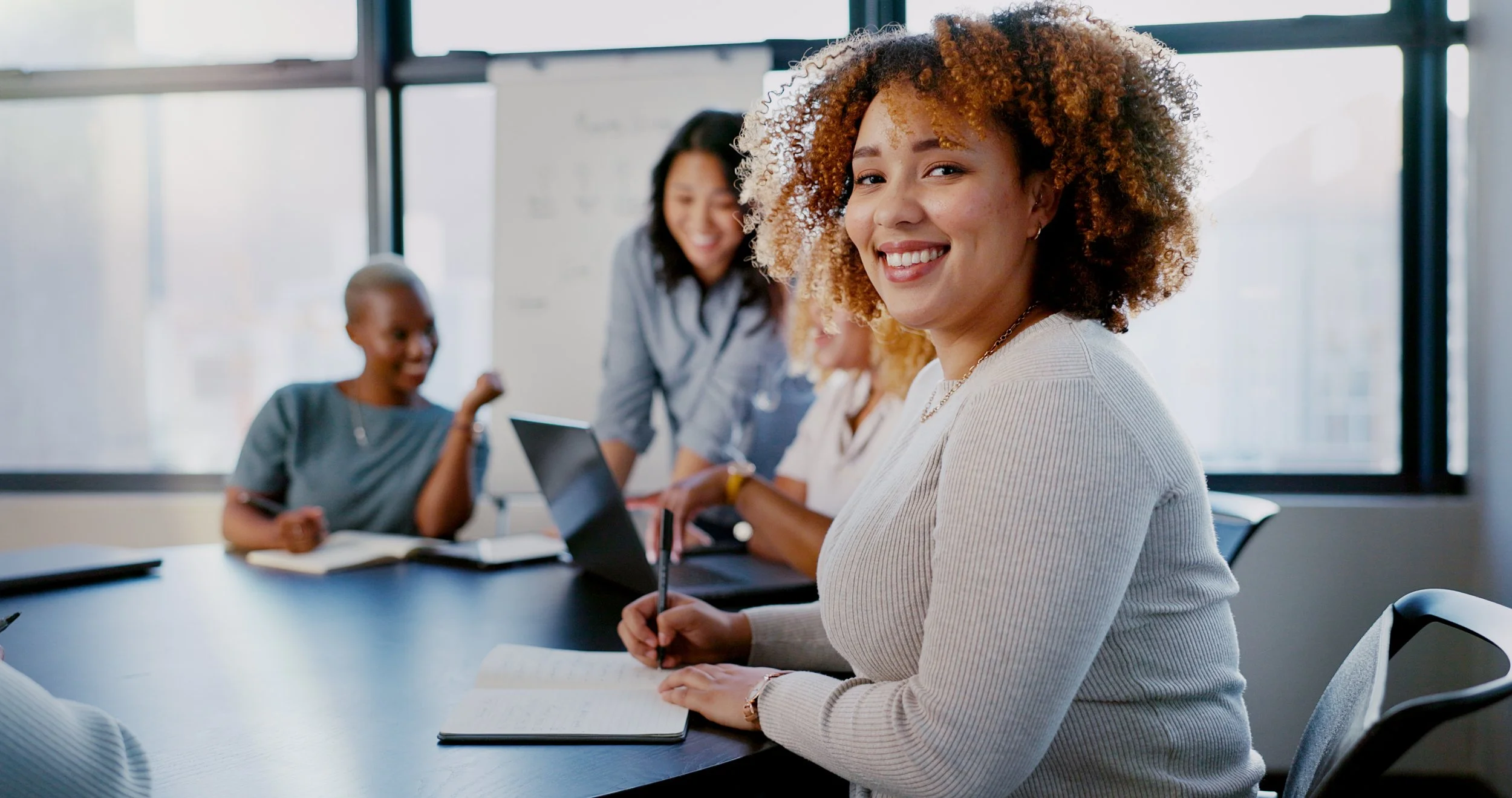 Smiling woman with curly hair sitting at a conference table with open notebook, in a meeting room with large windows, colleagues in background, some using laptops