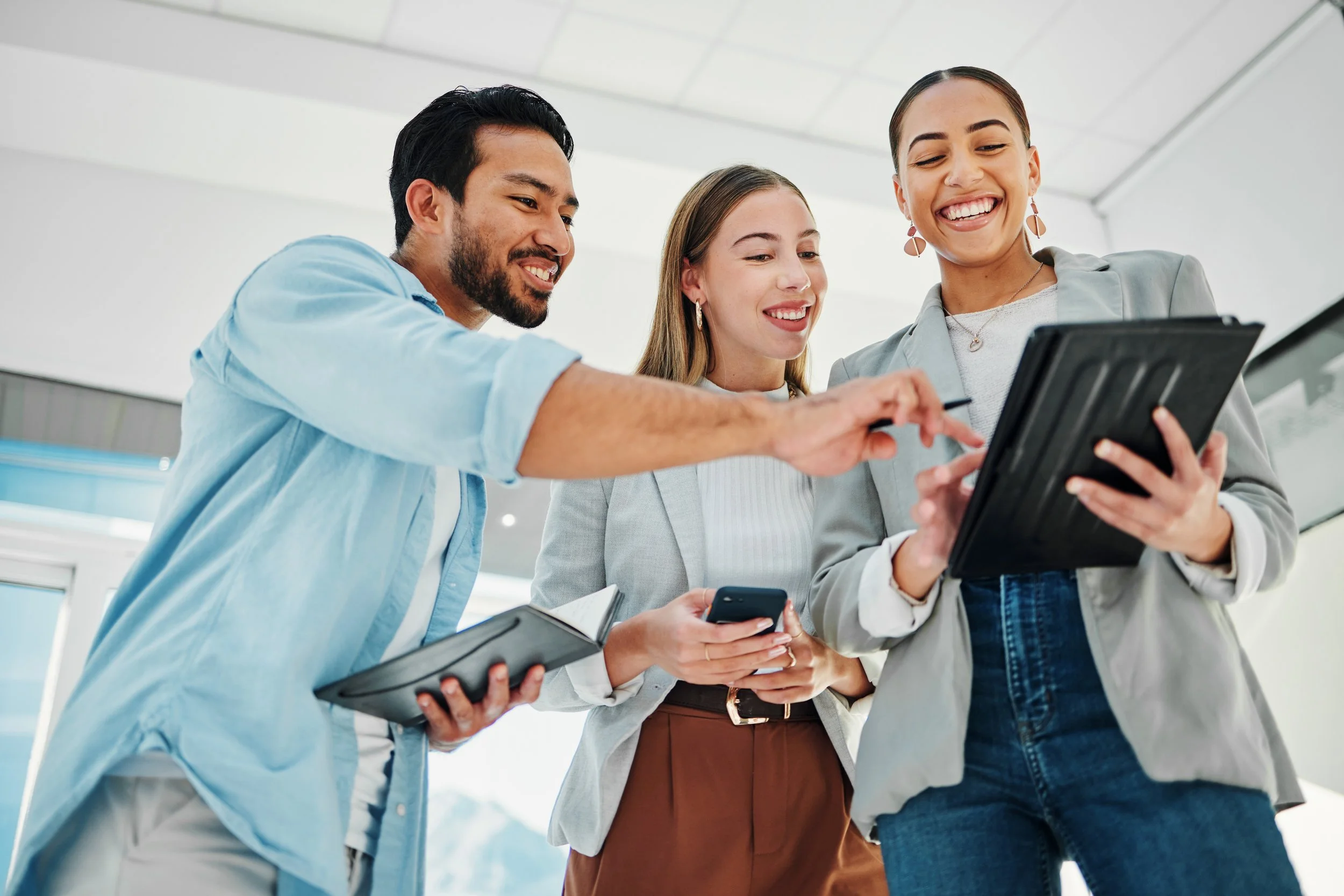 Three diverse business professionals looking at a tablet and smiling in an office setting.