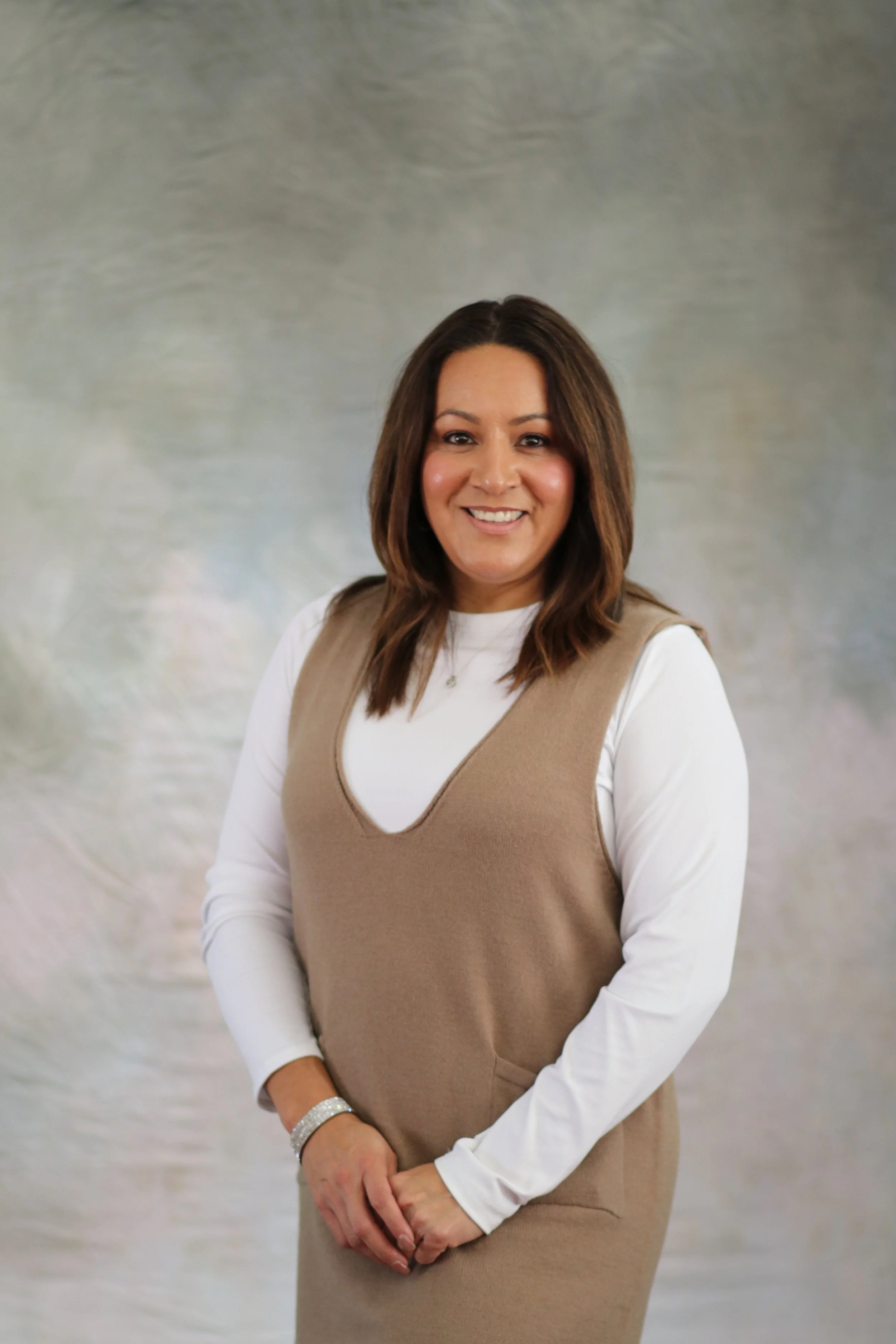 A woman with medium-length brown hair, smiling, wearing a white long-sleeve top with a beige sleeveless dress over it, standing against a gray textured background.