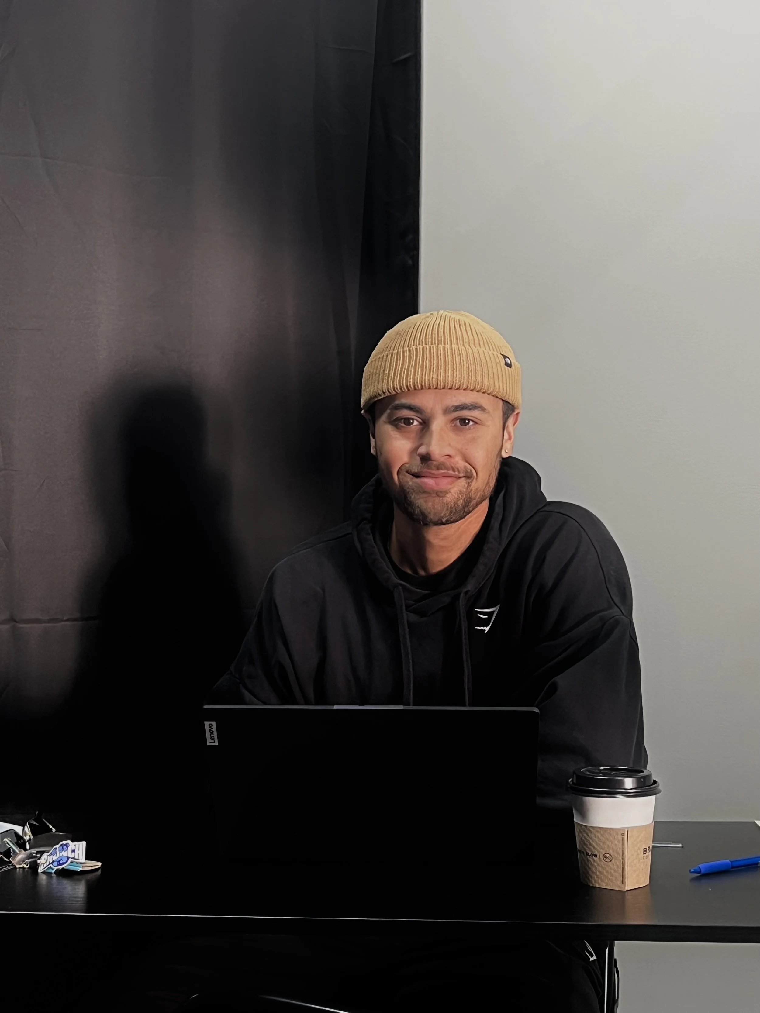 A man wearing a beige beanie and a black hoodie sitting at a desk with a laptop and a disposable coffee cup.