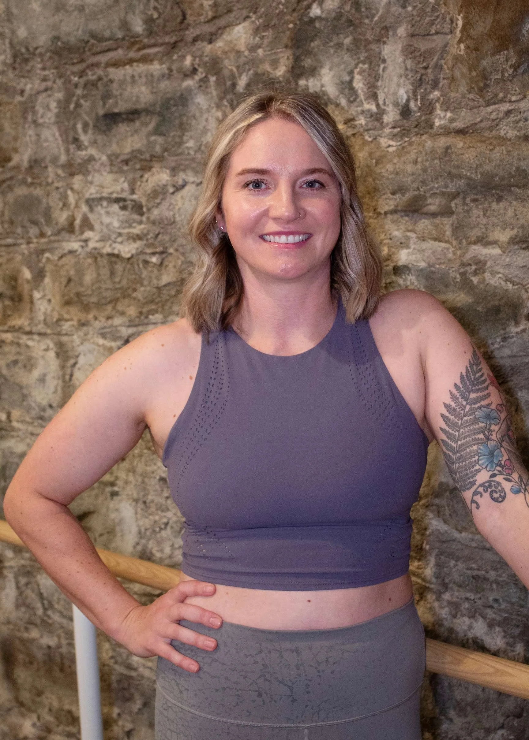 A smiling woman with light brown hair, blue eyes, and a nose piercing, standing outdoors with green foliage in the background.