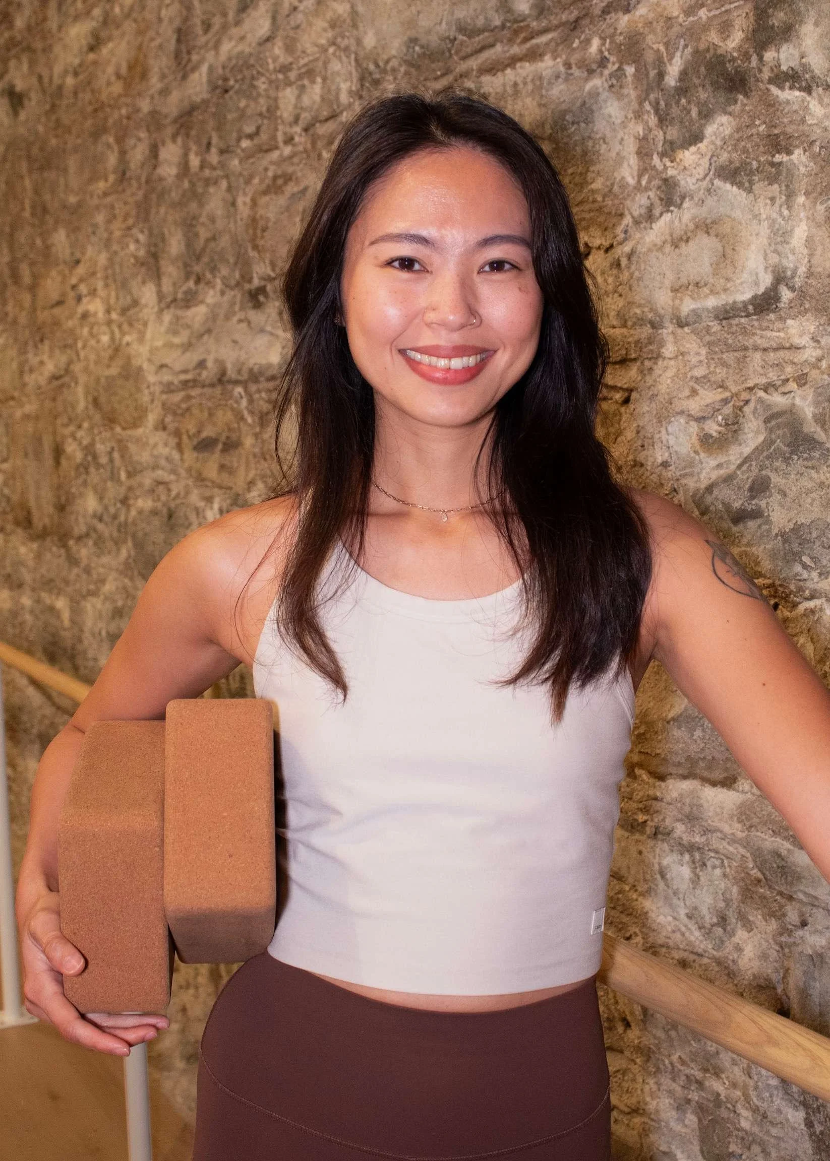 Person smiling while leaning on a reception desk in a yoga studio, wearing a black tank top and leggings; clothing rack with sportswear in the background.