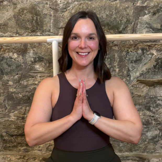 A woman sitting cross-legged on a yoga mat with hands in a prayer position against a stone wall backdrop.