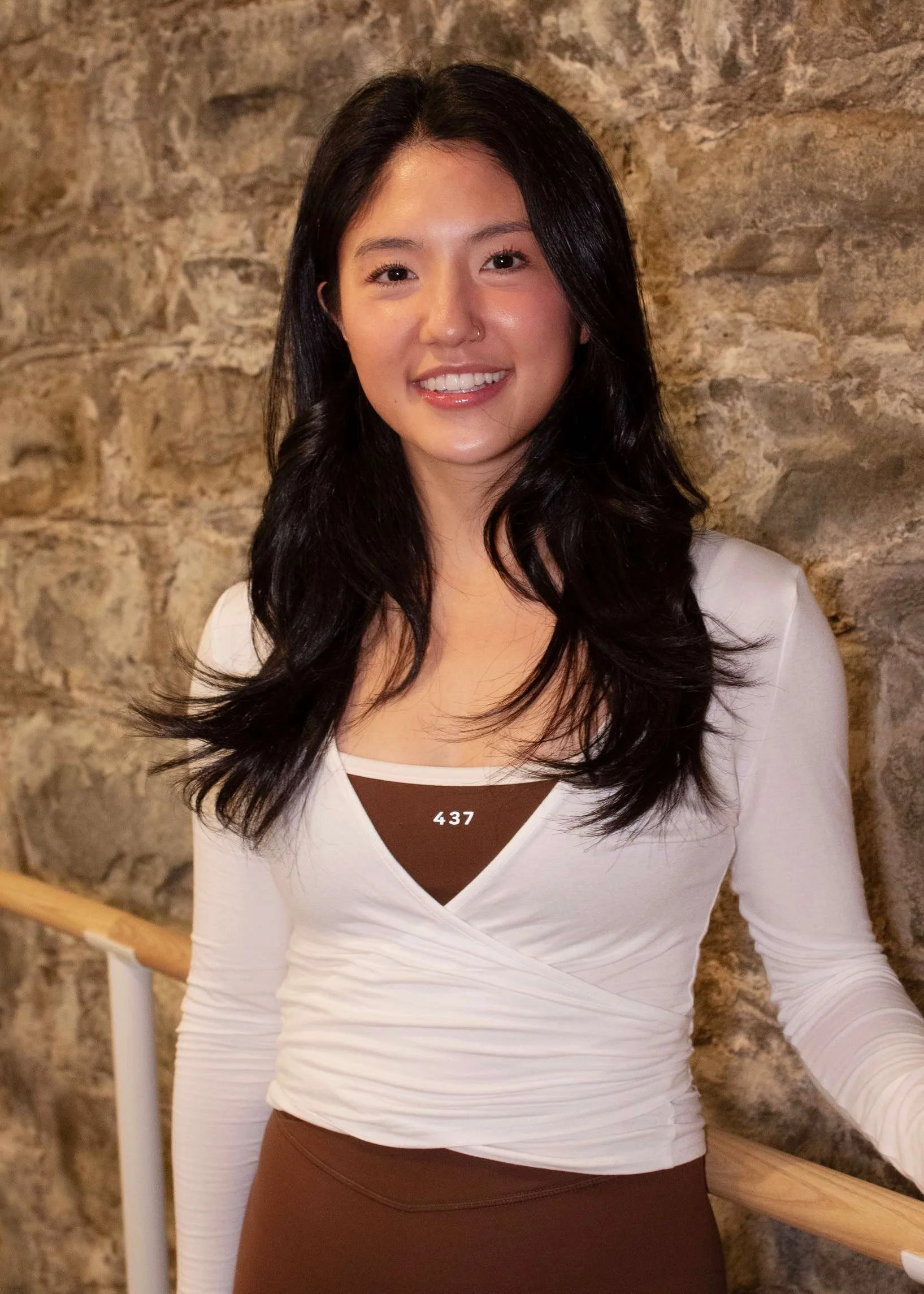 A young woman in workout attire holding a beige exercise ball in front of a stone wall in a fitness studio.