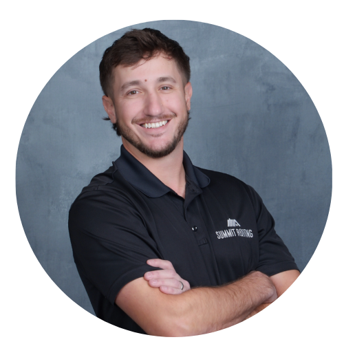 Smiling man with short brown hair, beard, and mustache, wearing a black polo shirt with logo and text, crossing arms, standing against a gray background.