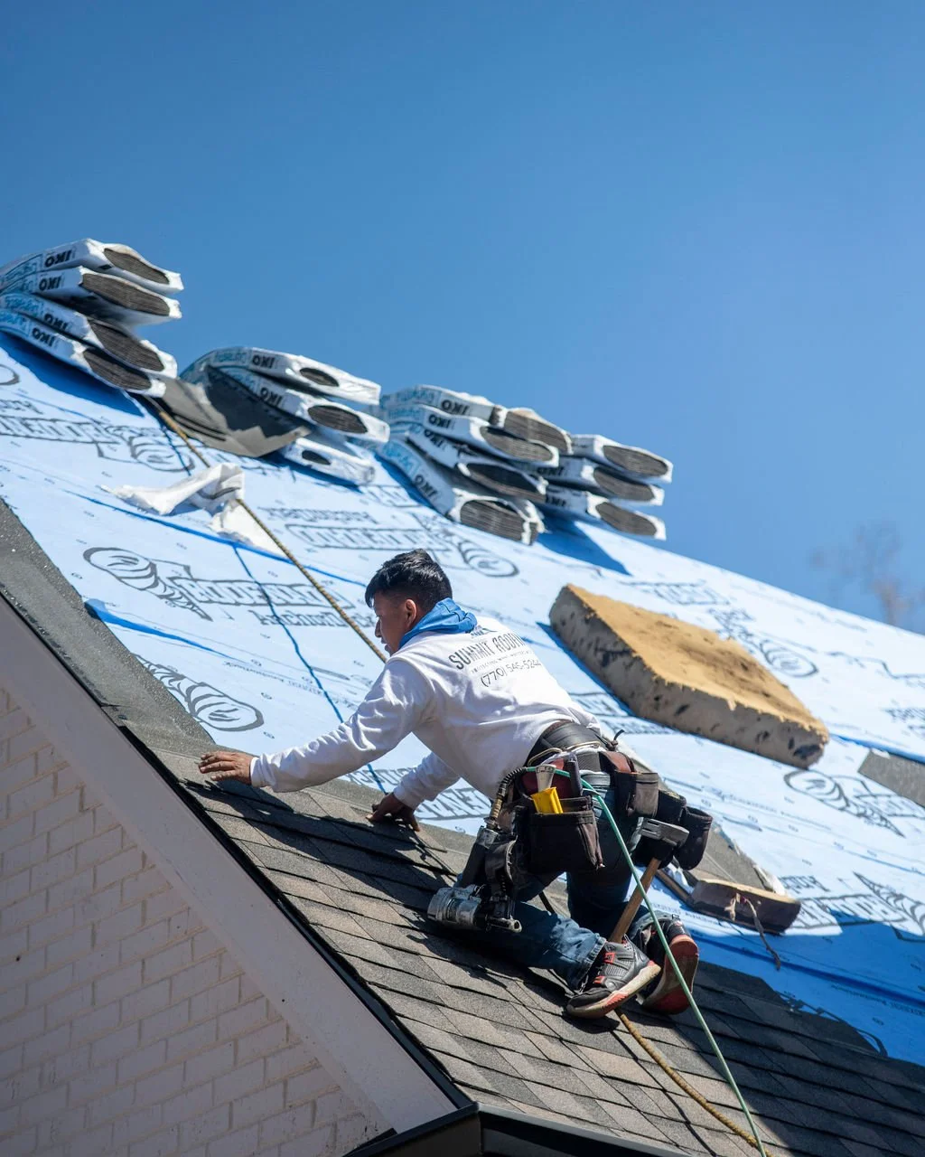 A construction worker installing roofing shingles on a house, with insulation and roofing materials visible above him.