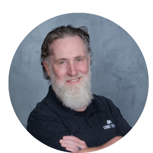 A smiling middle-aged man with a full beard and mustache, wearing a dark polo shirt, standing against a plain gray background.