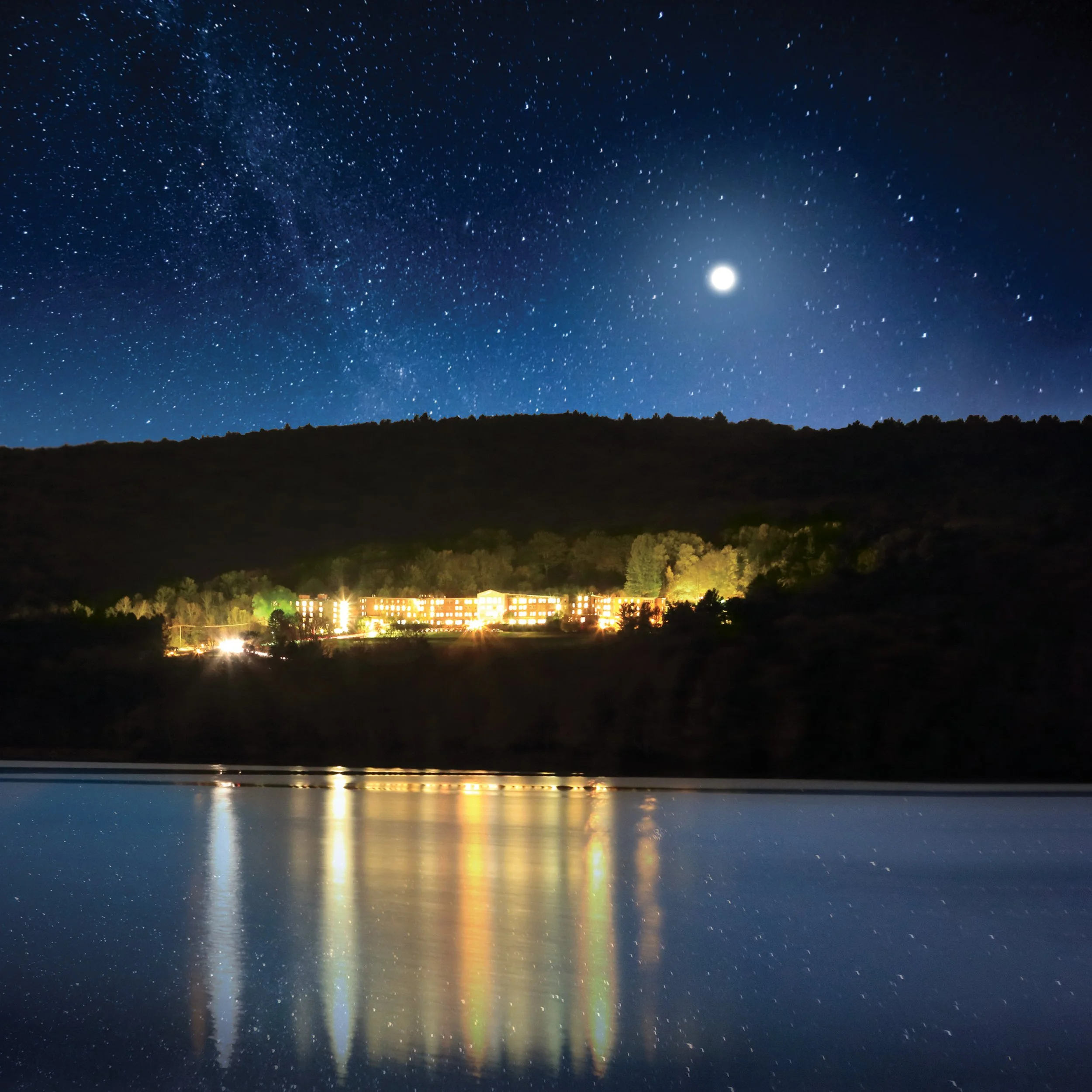 Night scene with starry sky and bright moon, illuminated building on hill, calm water reflecting lights.