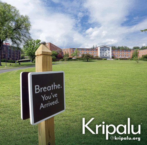 A sign on a wooden post in a grassy area reads, 'Breathe. You've Arrived.' with buildings and trees in the background on a partly cloudy day.