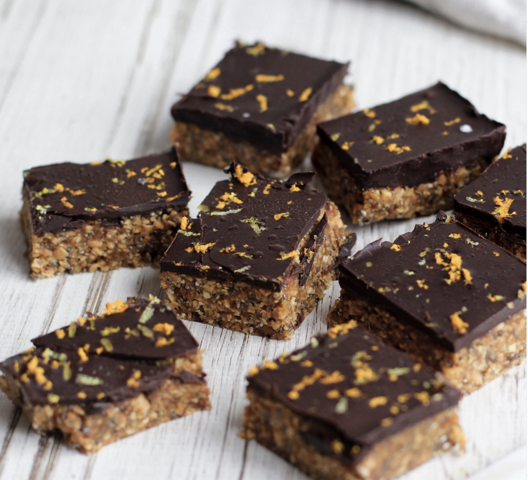 Close-up of multiple chocolate and nut bars on a white wooden surface, garnished with orange and green zest.