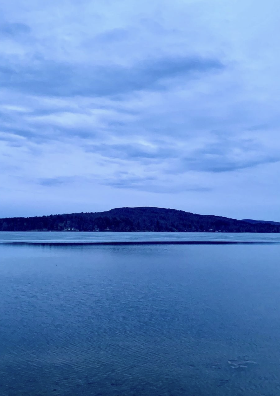 A calm lake with a tree-covered hill in the background under a cloudy sky.
