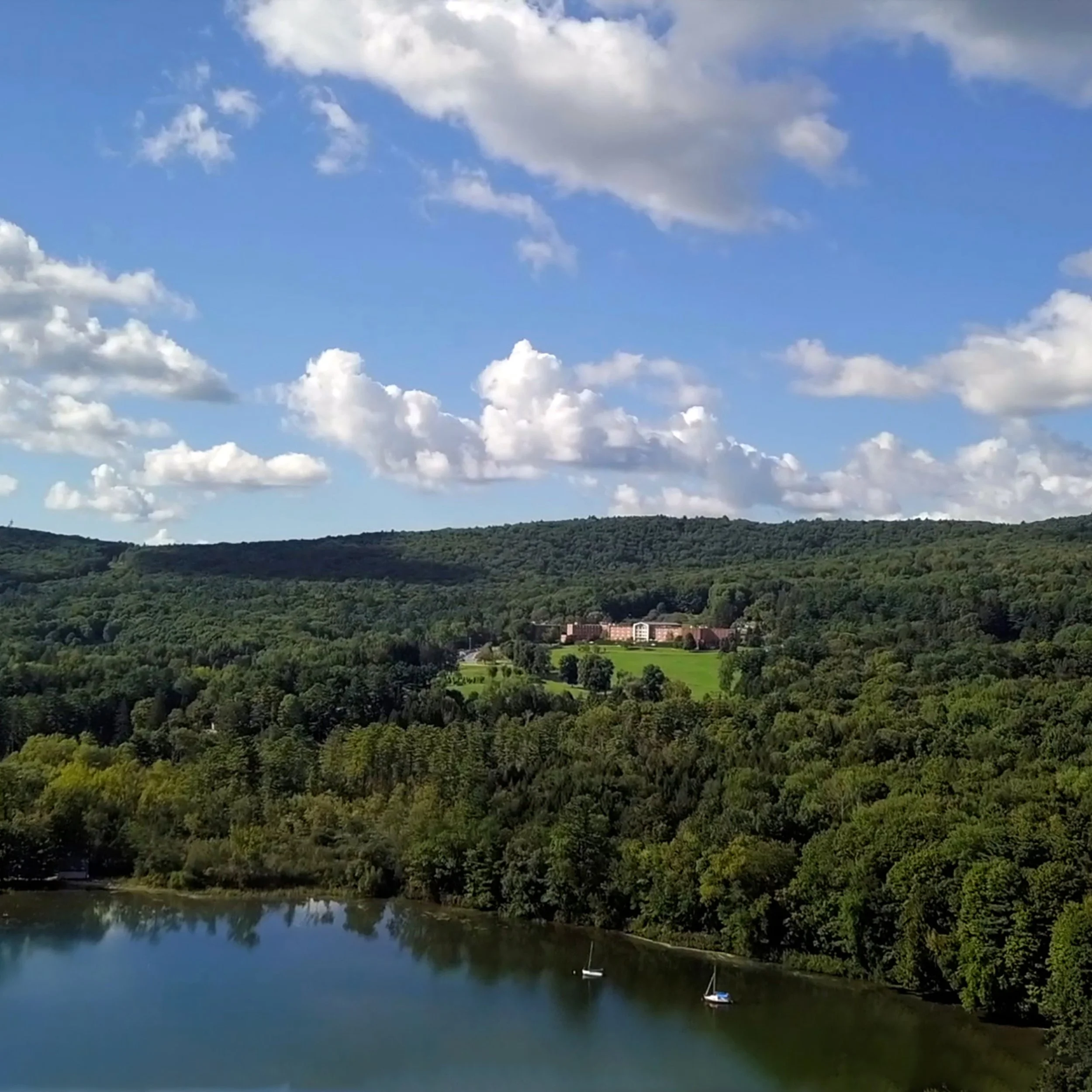 Scenic view of a body of water with two sailboats, surrounded by dense green trees, rolling hills, and a large building complex in the distance under a blue sky with scattered white clouds.