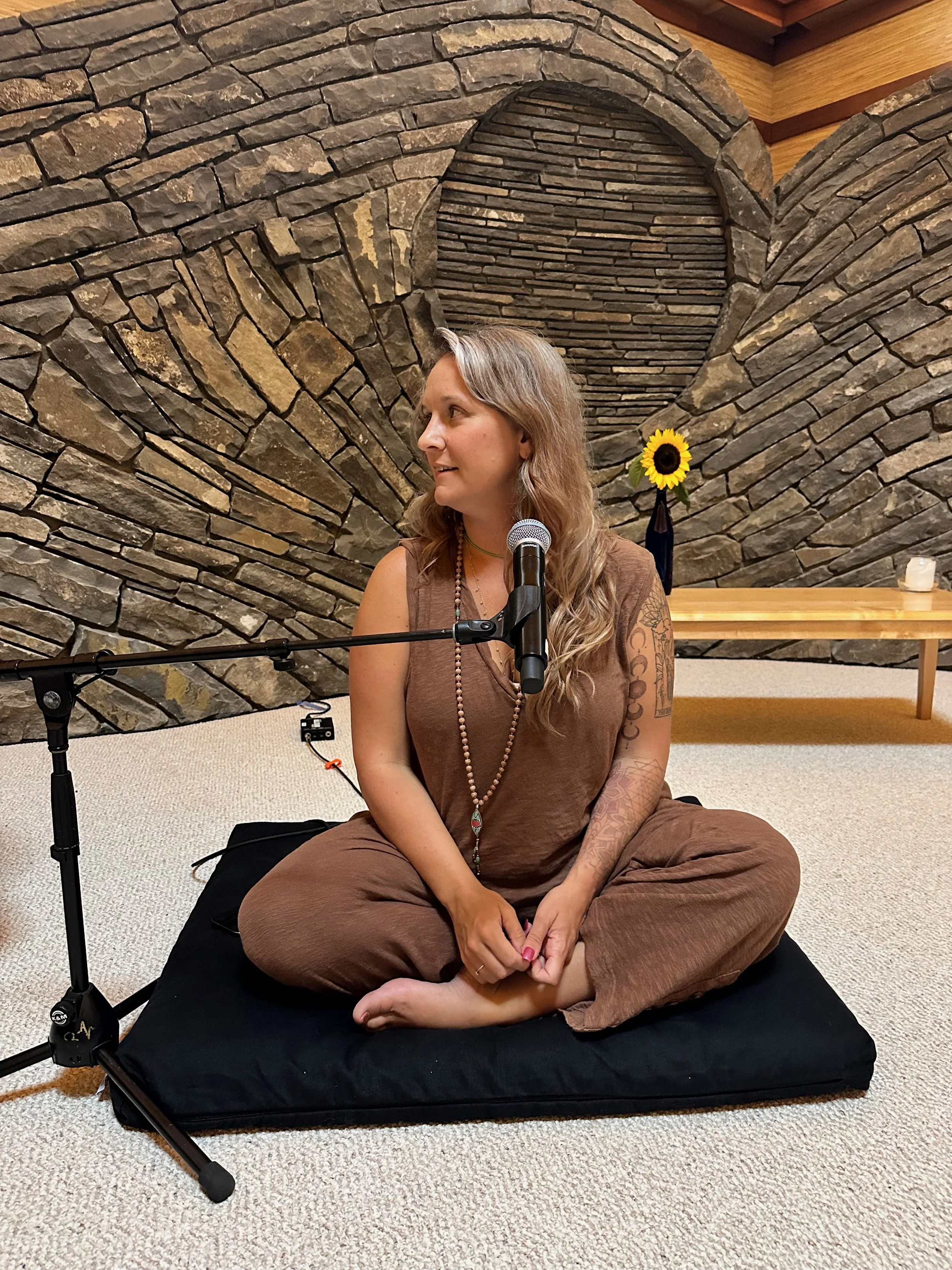 A woman with long wavy hair sitting on a black cushion on a beige carpet, wearing brown loose clothing, with tattoos on her arms, looking to her left, in front of a stone wall with a heart-shaped design, with a sunflower in a black vase on a wooden table behind her, and a microphone on a stand in front of her.