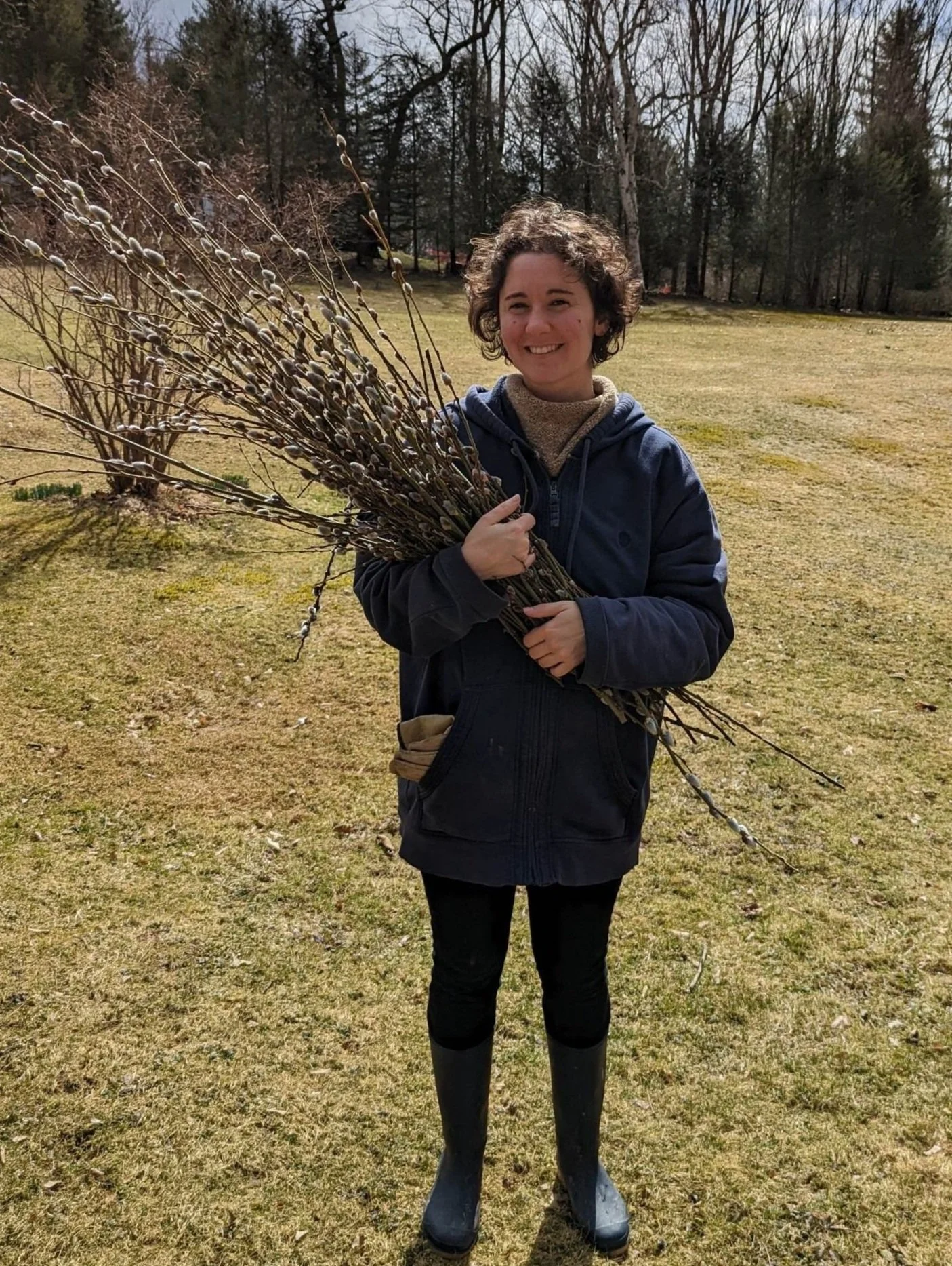A smiling woman holding a large bundle of pussy willow branches outdoors on a grassy lawn.