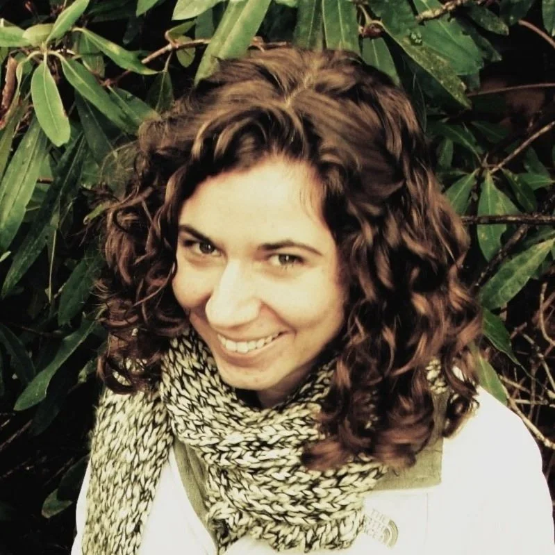 A woman with curly brown hair smiling, standing in front of green leafy plants, wearing a black and white scarf.