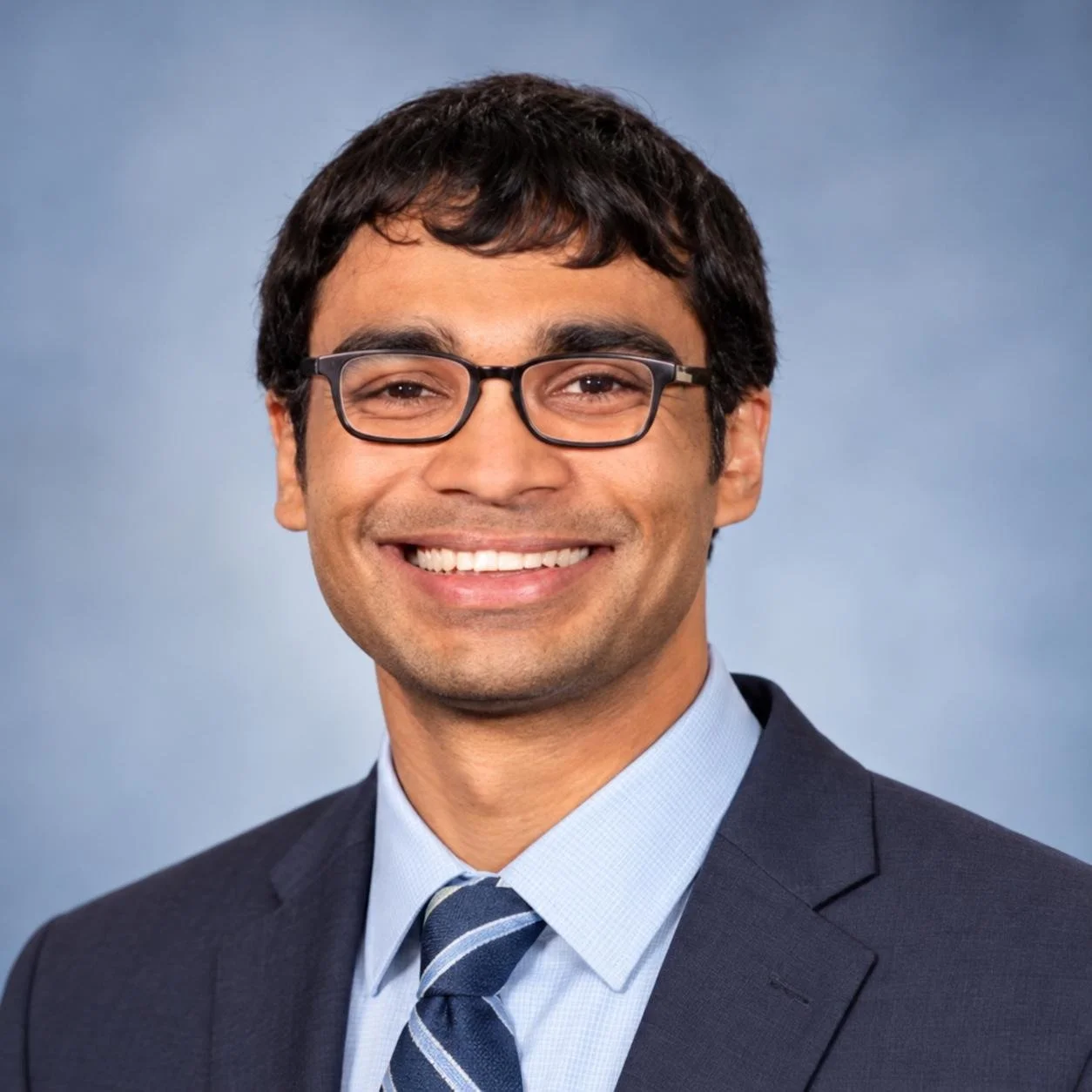 A smiling man with dark hair, glasses, wearing a suit and tie, against a blue background.