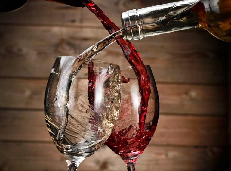 Red and white wine being poured into wine glasses, with ice in the glasses, against a wooden background.