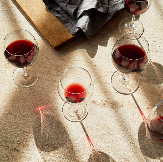 Five glasses of red wine on a light-colored surface with shadows, a wooden board, and a gray cloth nearby.