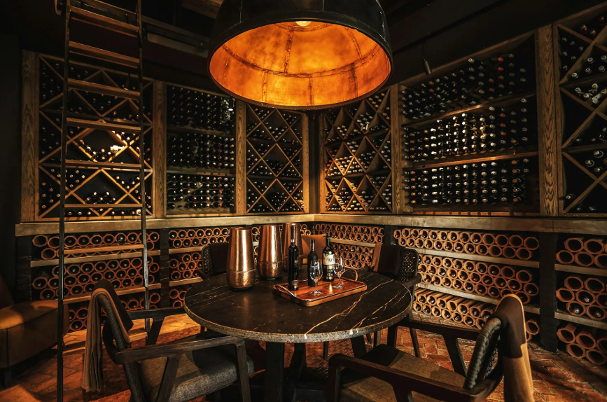 A cozy wine cellar with wooden shelves filled with wine bottles and a round table with wine bottles and metal carafes, illuminated by warm lighting.
