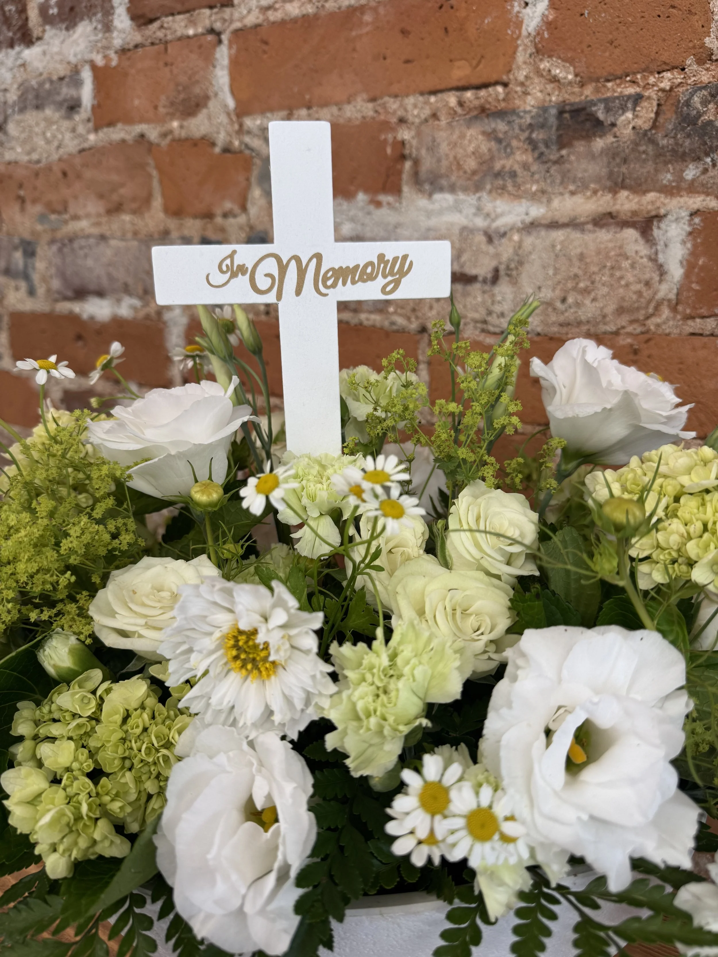 Close-up of the Garden of Remembrance sympathy arrangement with a keepsake memorial cross, white roses, lisianthus, hydrangea, and fresh greenery in a modern ceramic container
