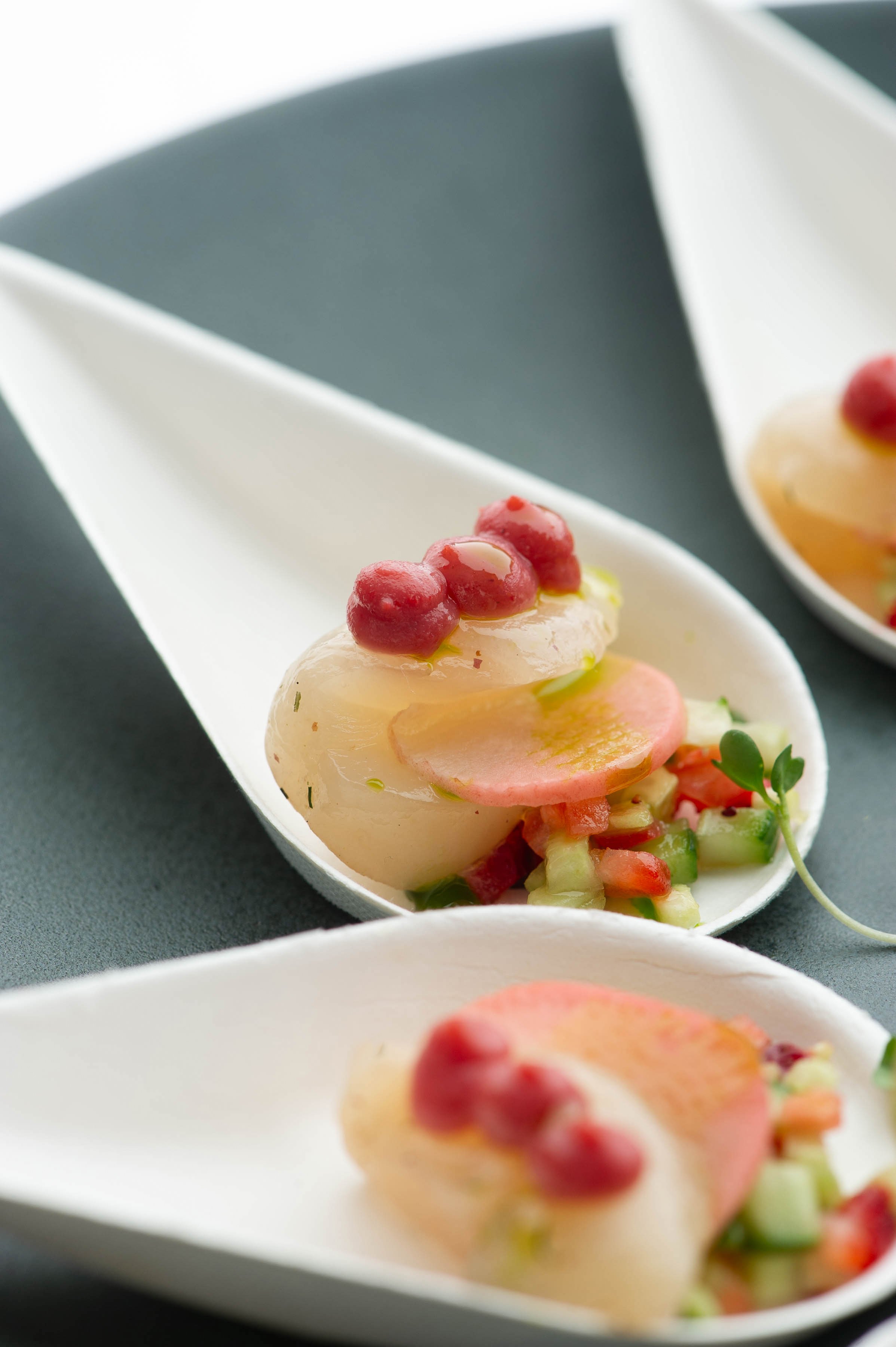 Close-up of white spoon holding a gourmet seafood dish with scallop, sliced radish, small red berries, and microgreens, with another spoon partially visible in the background.