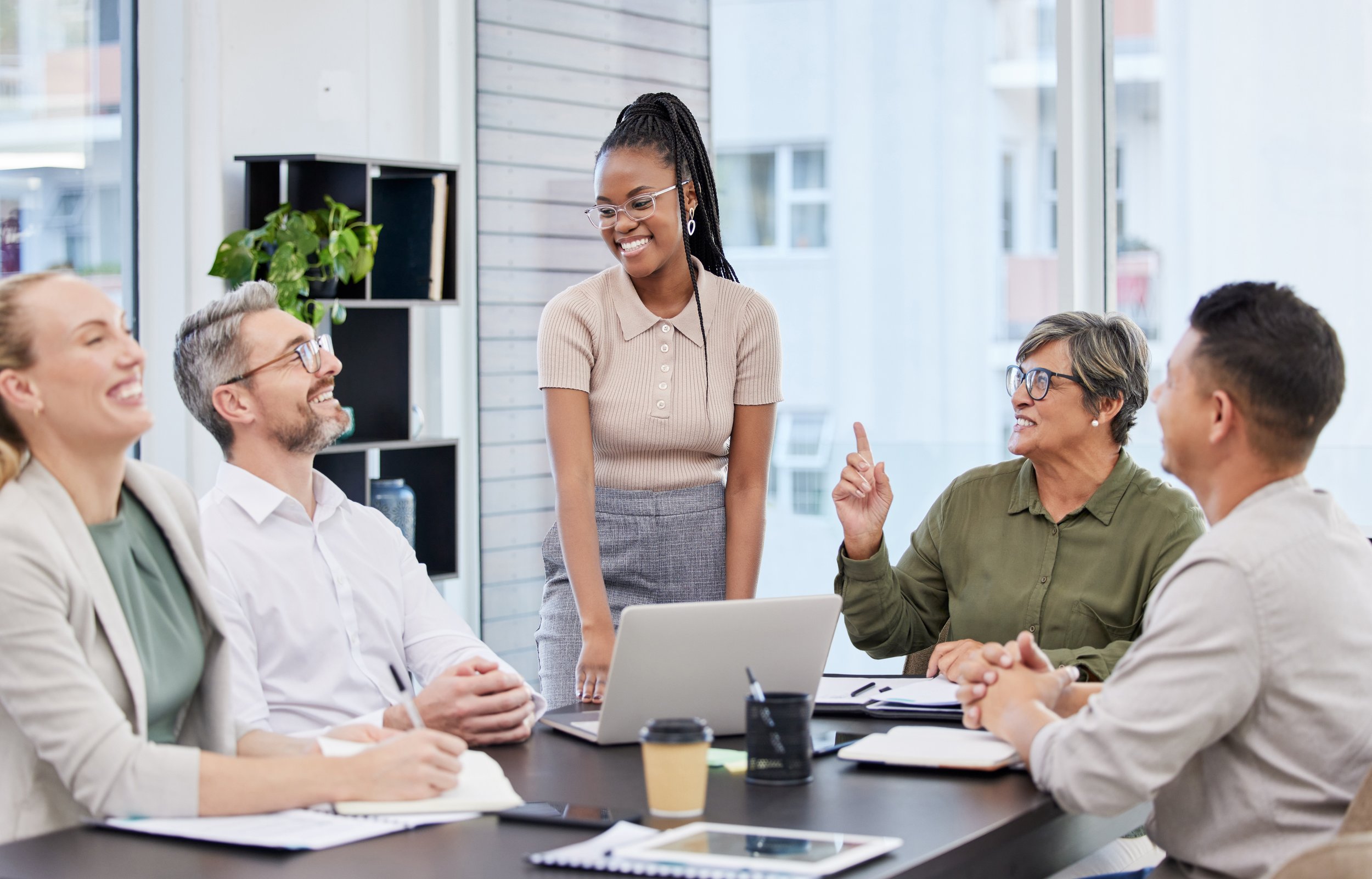 Diverse group of colleagues having a meeting in a modern office, with a woman standing and smiling, surrounded by three seated individuals, notebooks and a laptop on the table.