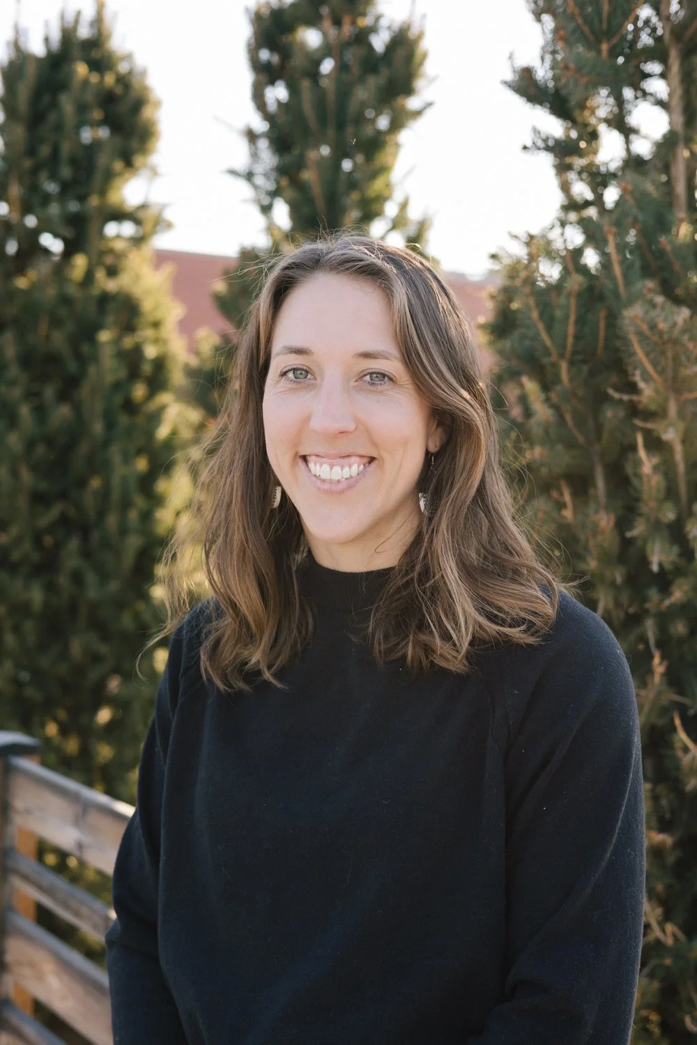 A woman with shoulder-length wavy brown hair and a black top outdoors, smiling with a background of trees and a building.