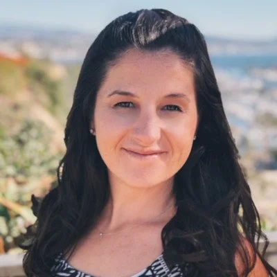 A woman with dark hair smiling outdoors with a scenic background of greenery and a body of water.