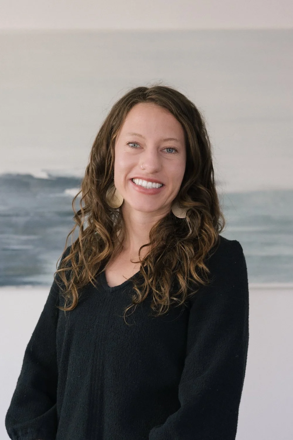 A woman with long, curly brown hair, light skin, and a nose ring, smiling and wearing a black sweater, standing in front of a beach scene painting.