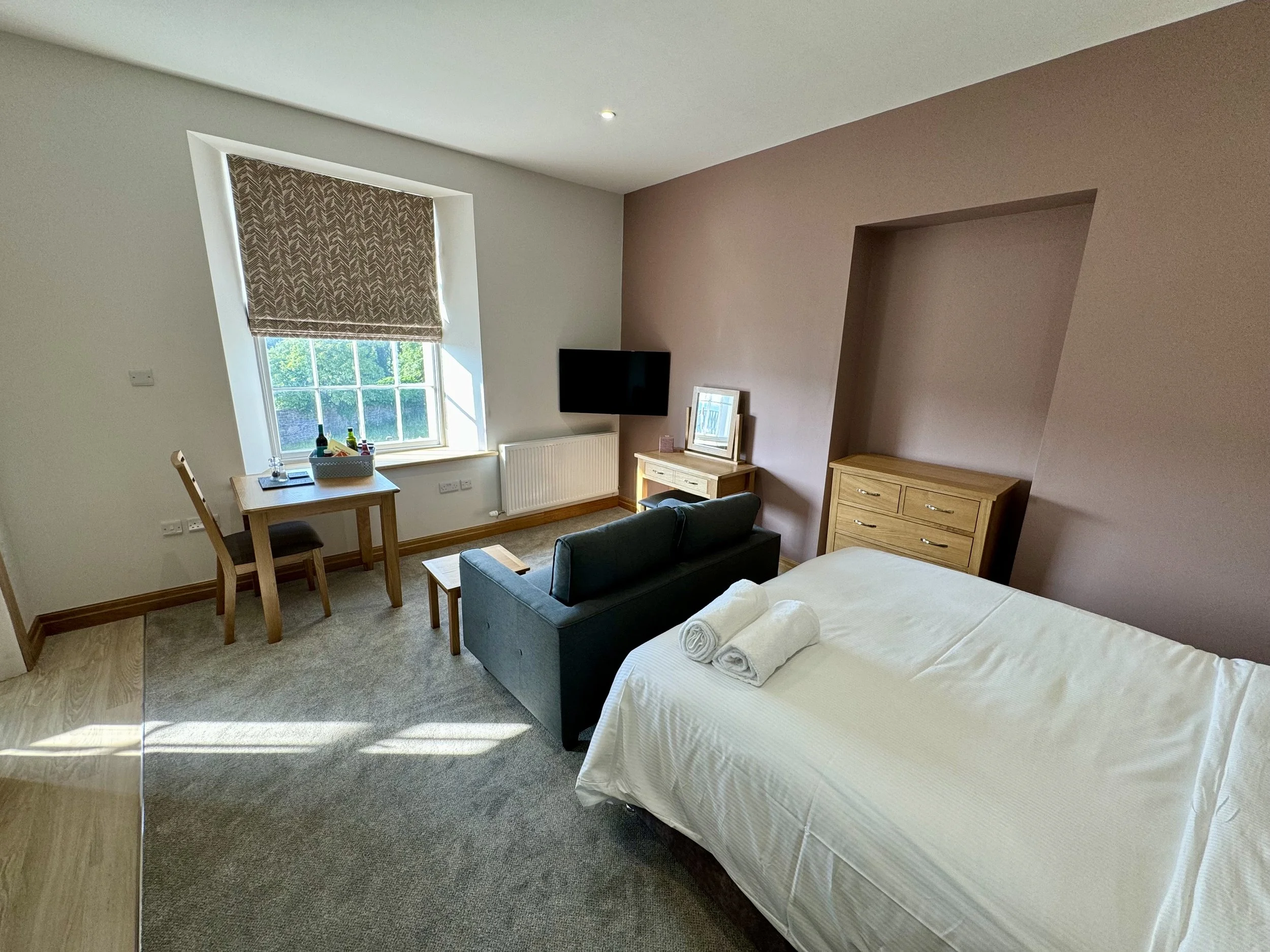 A hotel room with a bed, a small sofa, a wooden desk with a chair near a window, and a flat-screen TV mounted on the wall.