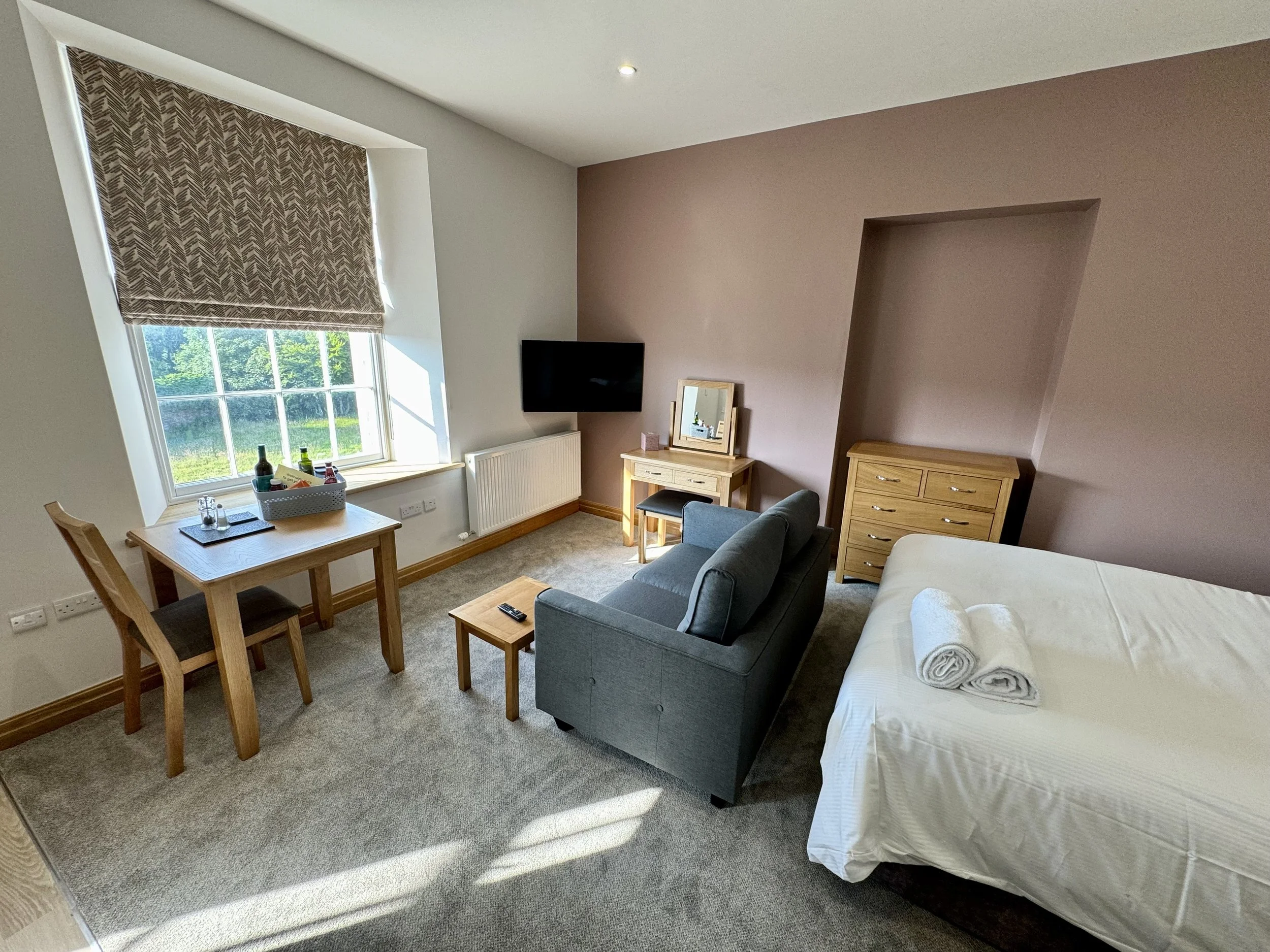 A hotel room with a bed, a sofa, a small table, a dining table with two chairs, a window with a patterned blind, a wall-mounted TV, a dresser, and a vanity with a mirror. The bed has two rolled towels on top.