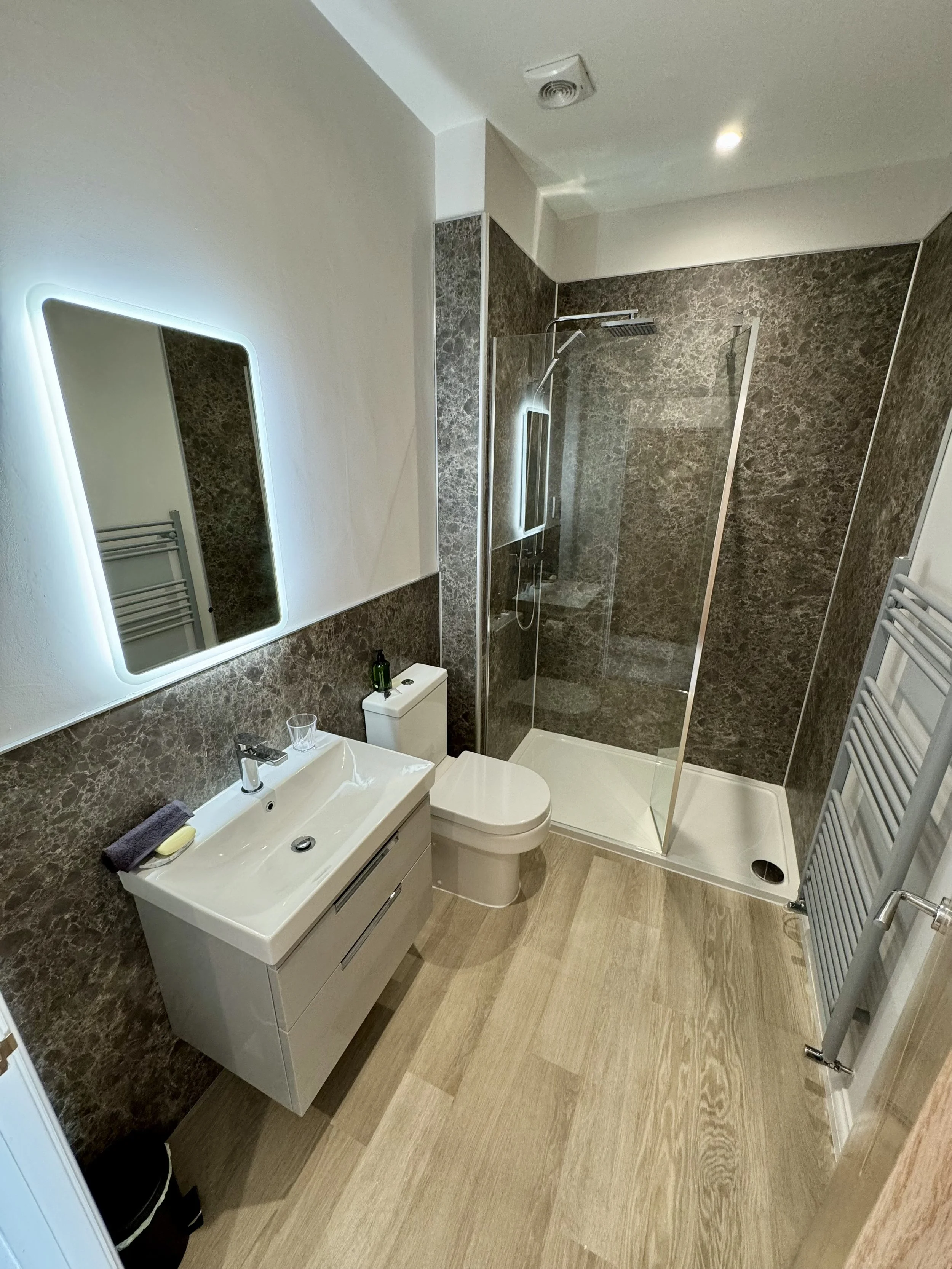 Modern bathroom with white sink vanity, wall-mounted oval mirror, toilet, walk-in shower with glass door, dark marble walls, light wood flooring, and heated towel rail.