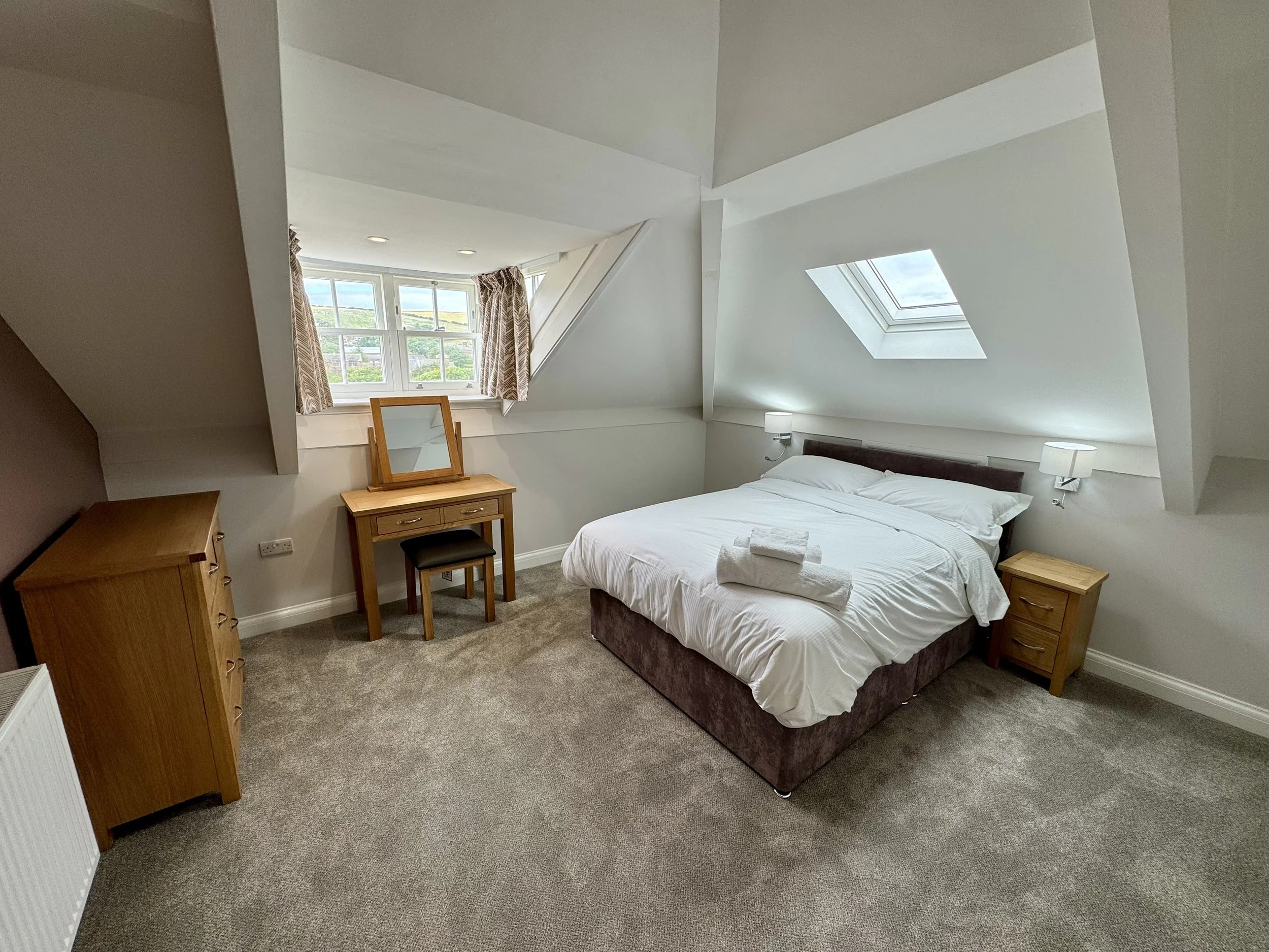 A cozy bedroom with a sloped ceiling, double bed with white linens and folded towels, two bedside lamps, a wooden dresser, and a dressing table with mirror. A window with curtains and a skylight are visible, offering natural light.