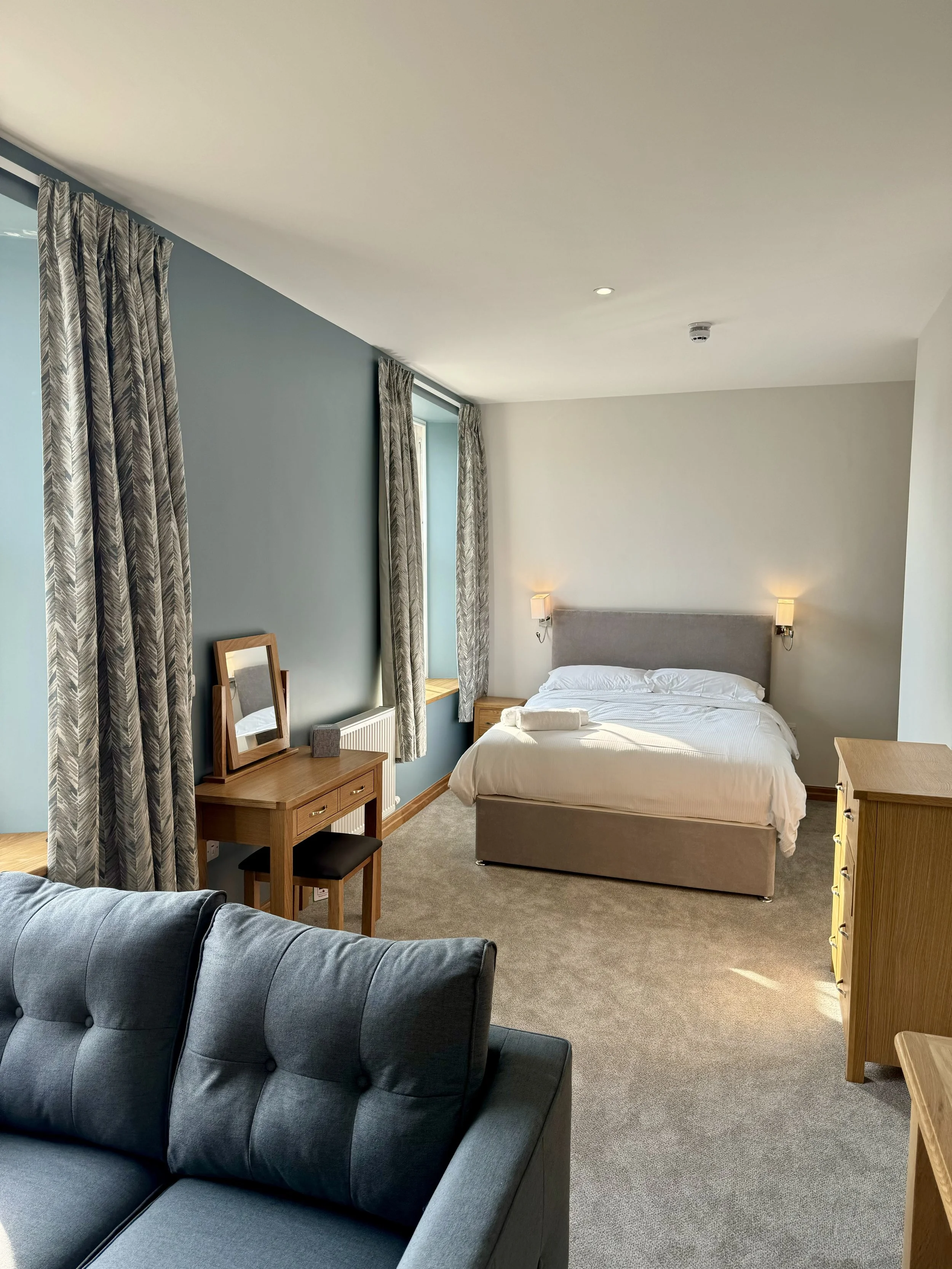 A hotel room with a bed, a dresser, a vanity table with a mirror, and a sofa, featuring light-colored walls, gray curtains, and natural lighting.