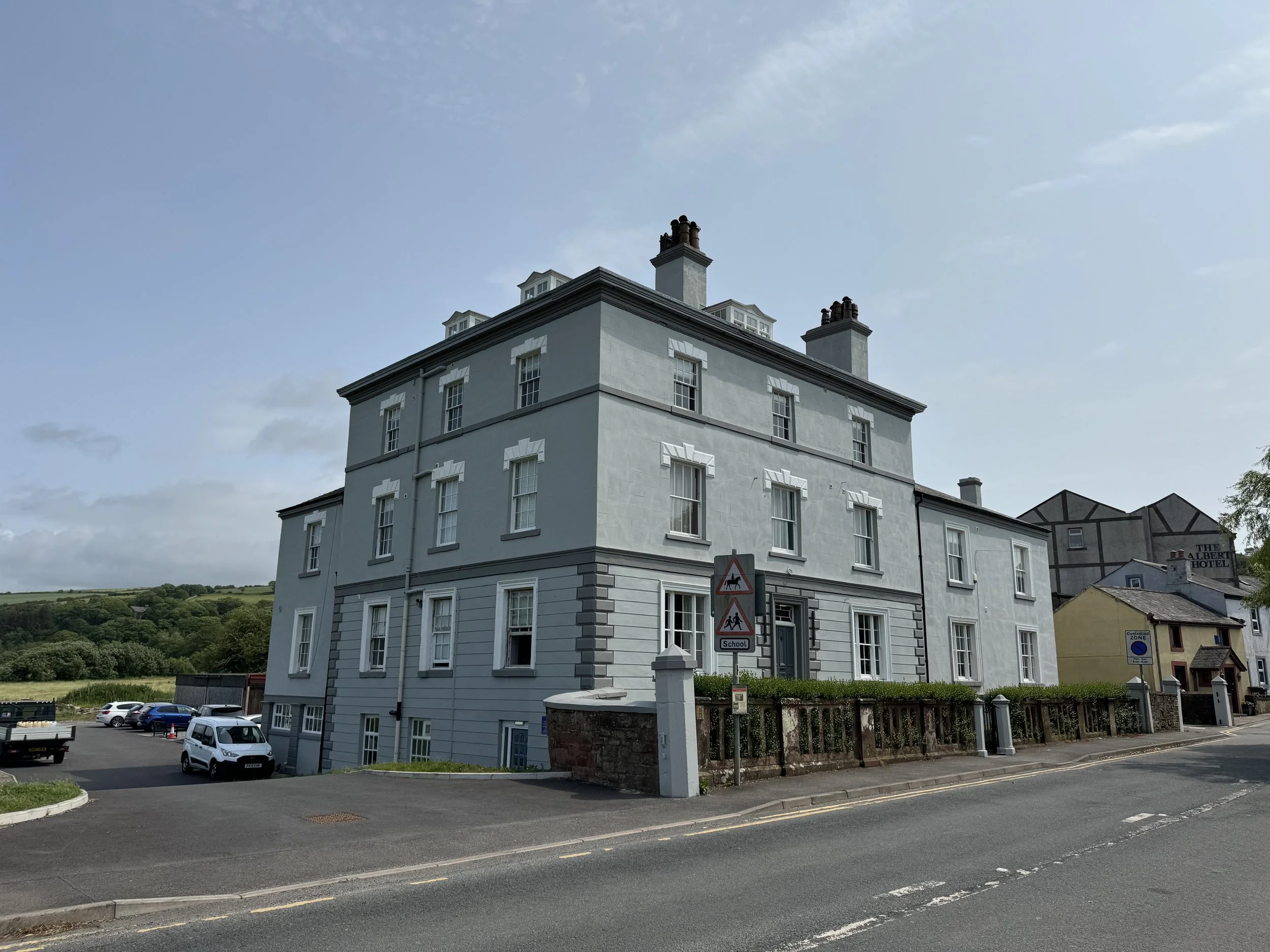 A large, four-story gray building with white trim and multiple windows situated on a street corner. There are cars parked nearby and several traffic and pedestrian signs in front of the building. The sky is partly cloudy with a green hillside in the 