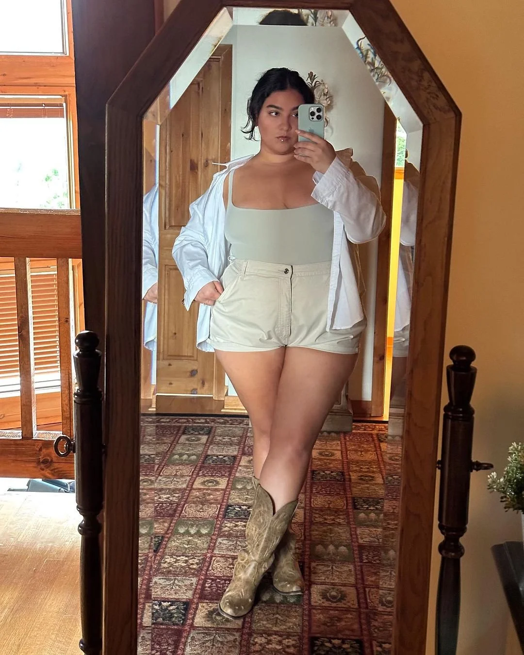 Nadia is taking a mirror selfie in a cabin house wearing skims bodysuit, white oversize shirt, and khaki high waisted shorts.
