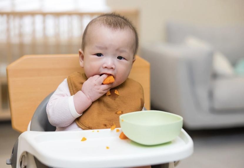 image of an infant sitting in a high chair eating lightly steamed carrots.