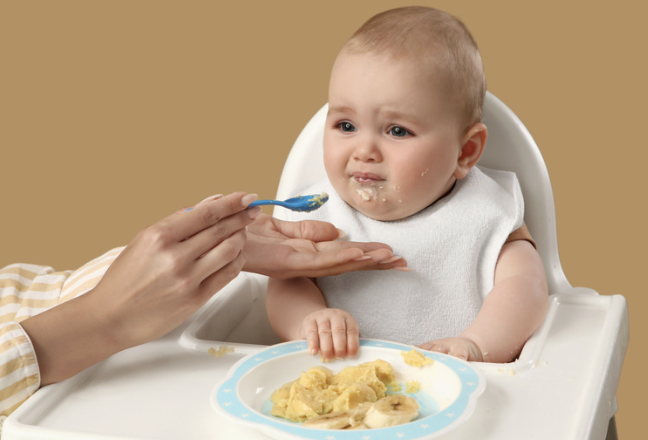 image of a young baby eating in a high chair with an unhappy look in his/her face.