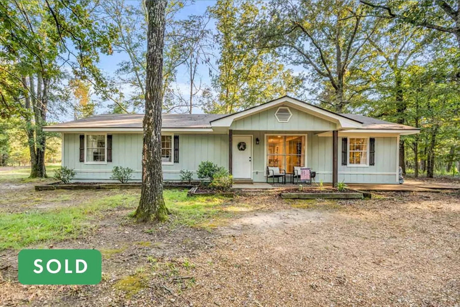 Single-story house with light blue exterior and dark shutters, front porch with seating, surrounded by trees, with a gravel driveway in front.