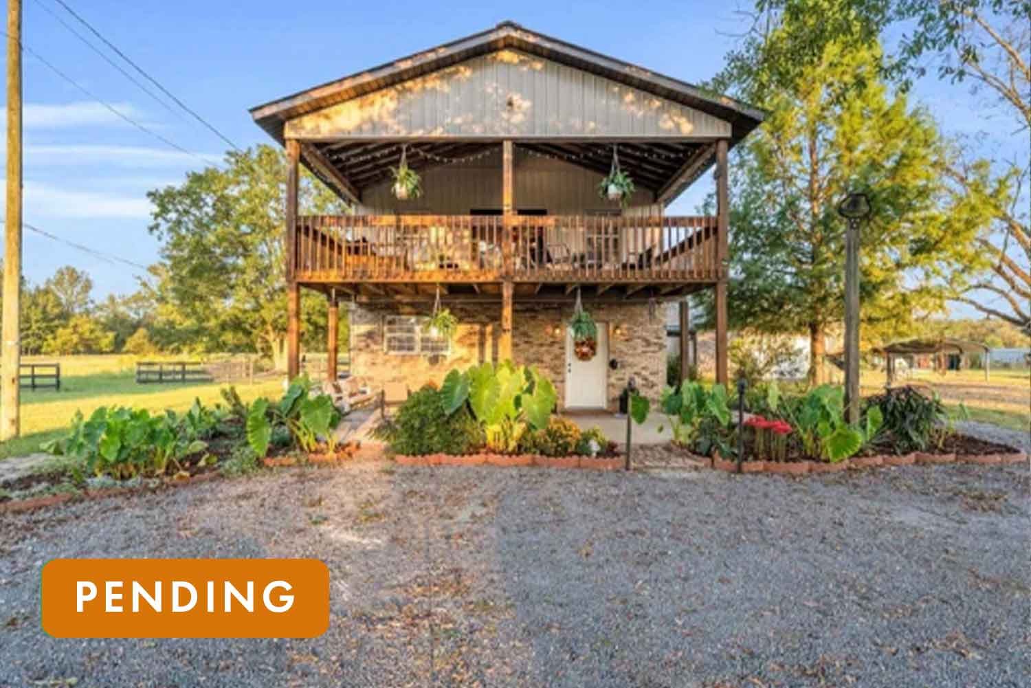 A two-story house with a wooden balcony on the upper level and brick exterior on the lower level, surrounded by a garden with large green leaves and plants, a gravel driveway, and trees in the background.