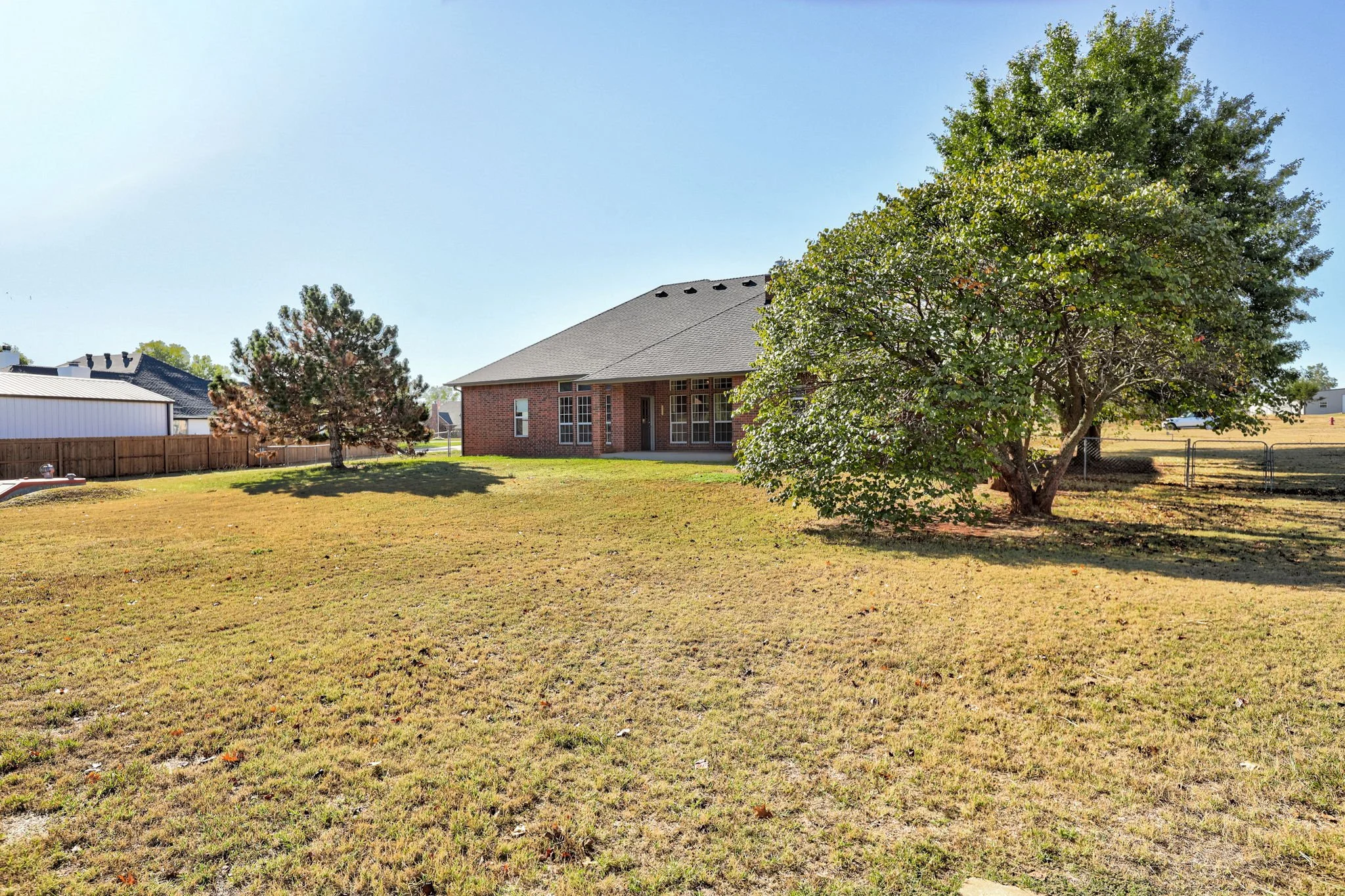 A backyard with a grassy lawn, multiple trees, a brick house with large windows, and a wooden fence under a clear blue sky.