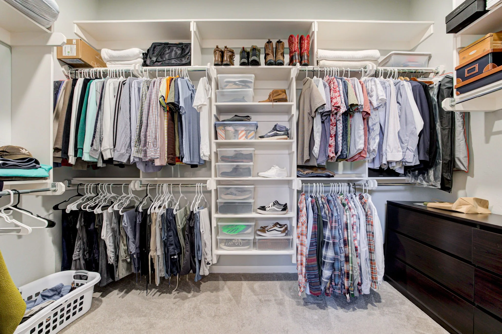 A walk-in closet featuring neatly organized rows of hanging clothes, plastic storage bins with shoes and accessories, a dark wooden dresser, and shelves holding shoes and boots.