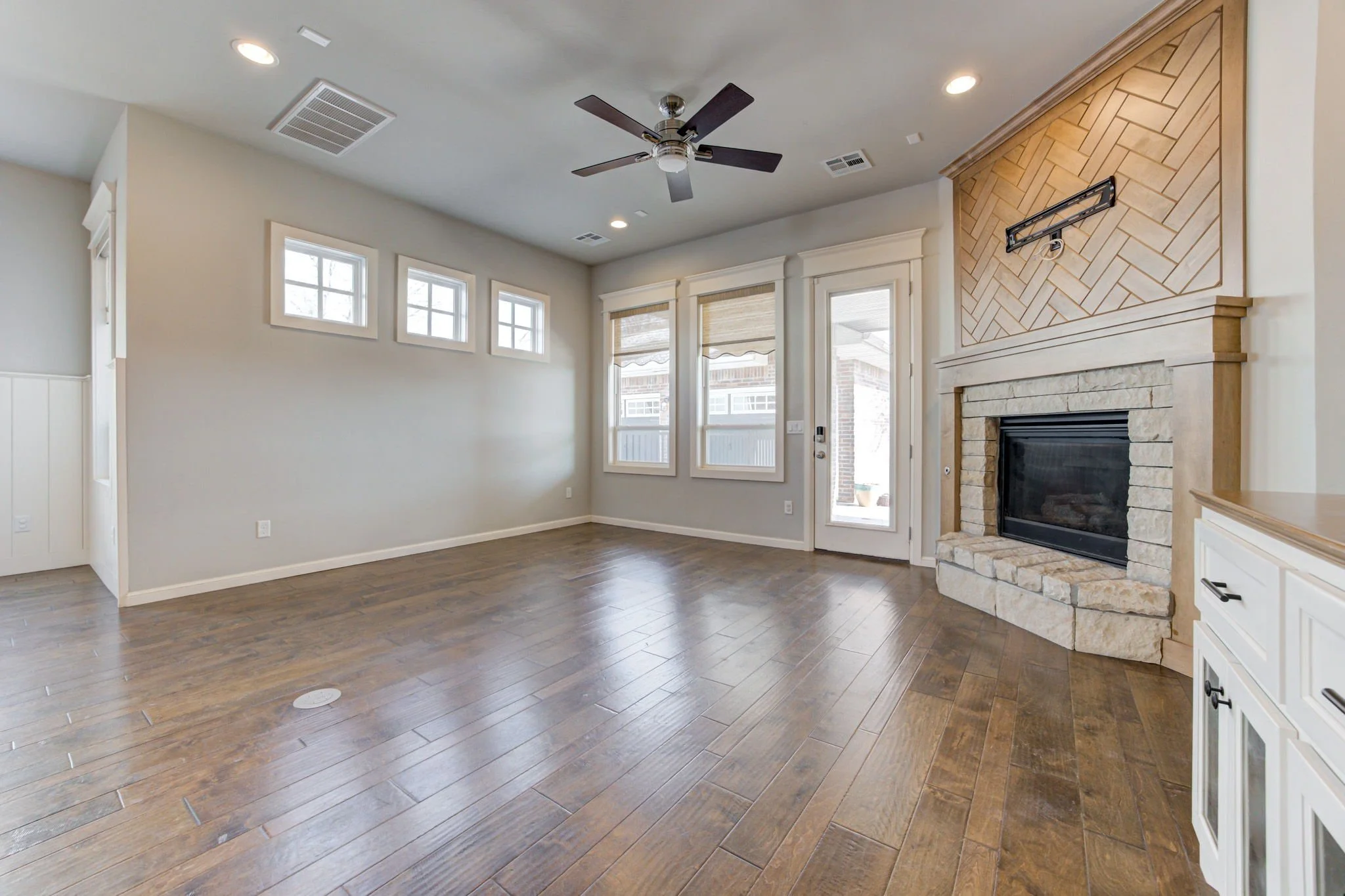 Empty living room with hardwood floors, a ceiling fan, a stone fireplace, four rectangular windows, a glass door, and a small window above the door.