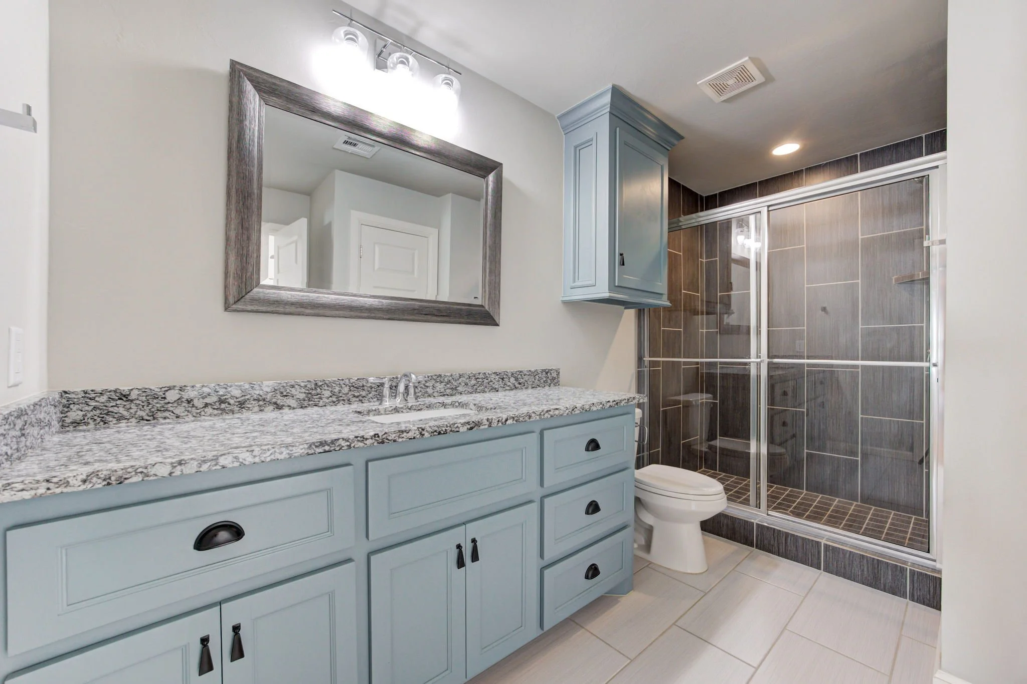 Bathroom with a blue vanity, granite countertop, mirror, shower with glass door and dark gray tile, and a toilet.