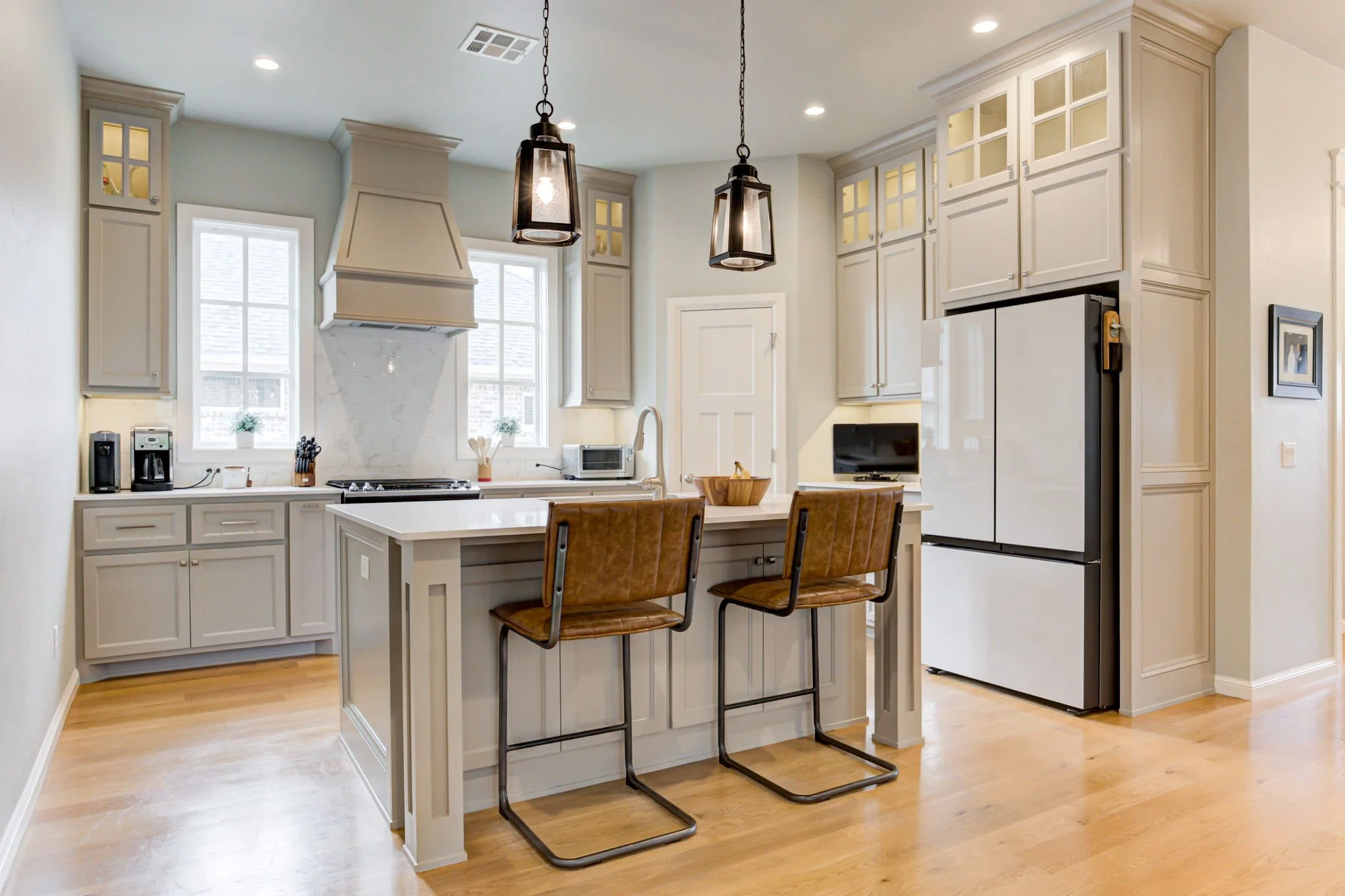 Bright, modern kitchen with white cabinets, two pendant lights, a central island with two brown leather bar stools, a refrigerator, and a window above the sink.