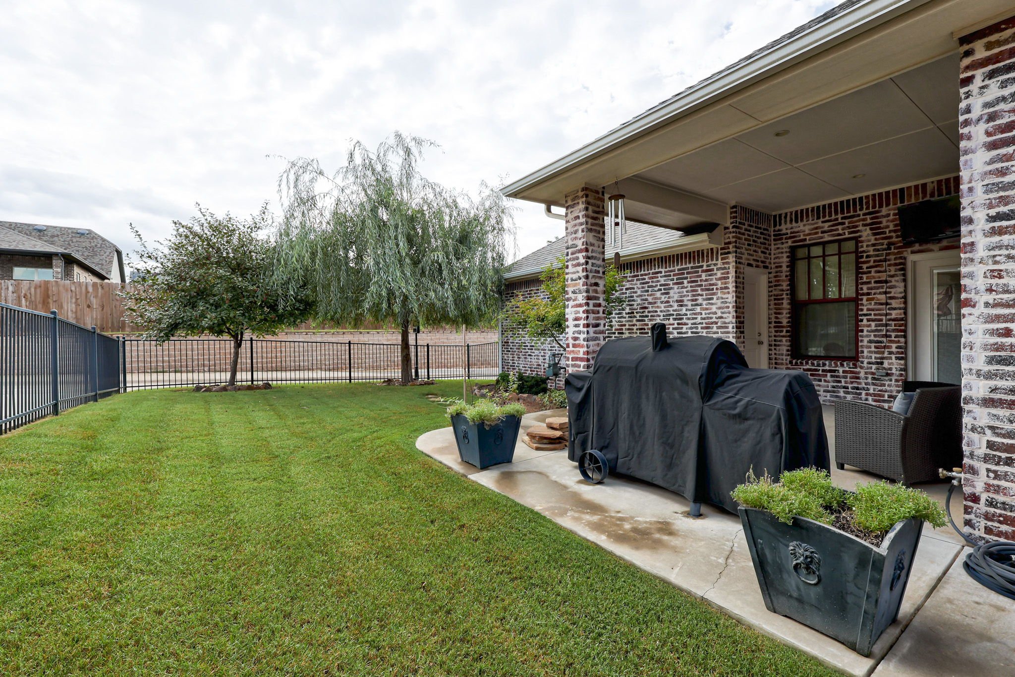 Backyard with green lawn, two trees, a covered grill, patio plants, and a fence