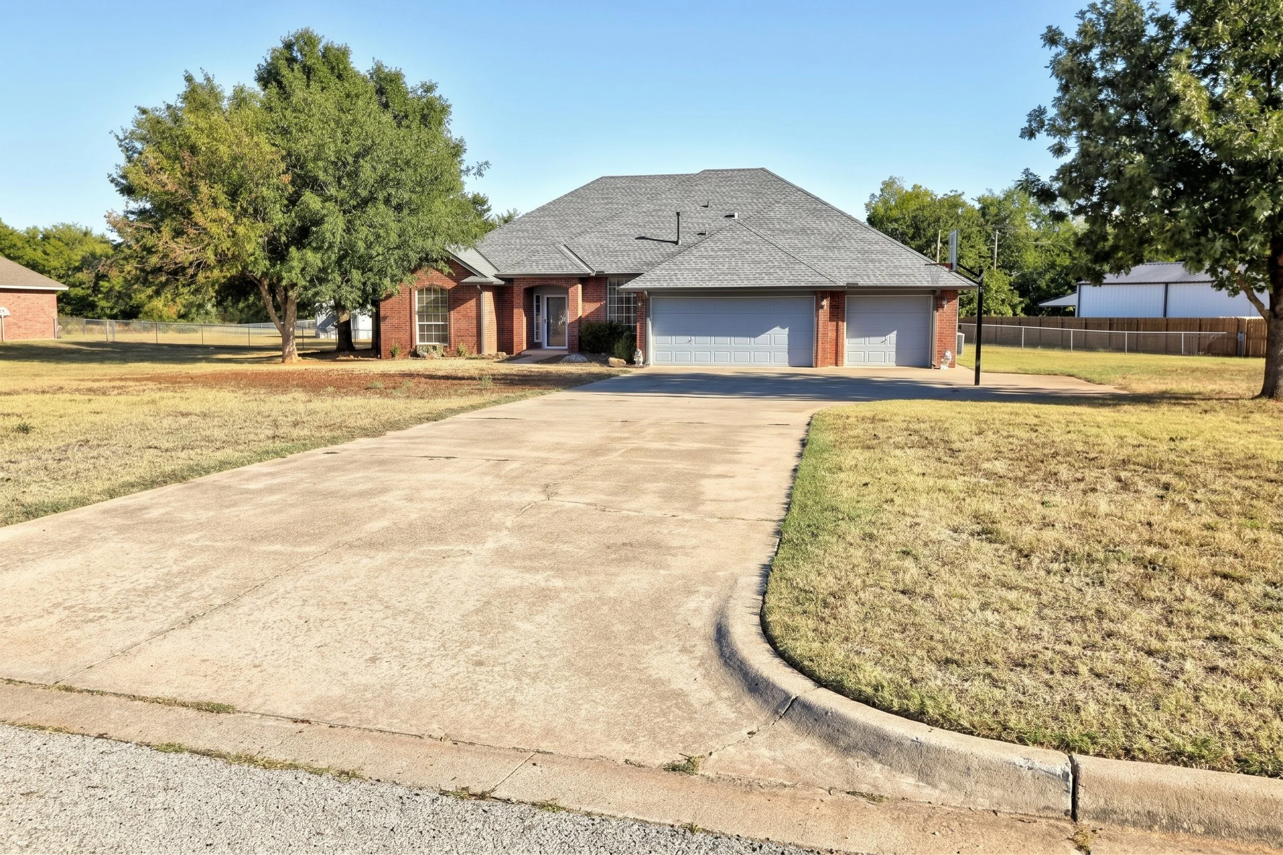 A suburban house with a brick exterior, gray shingle roof, two-car garage, and a driveway. There are large trees on the front yard, with green grass and a weathered concrete sidewalk leading to the house. The sky is clear and sunny.